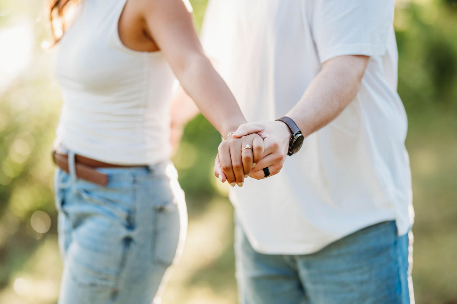A couple in casual attire holding hands outdoors with a soft bokeh background effect.