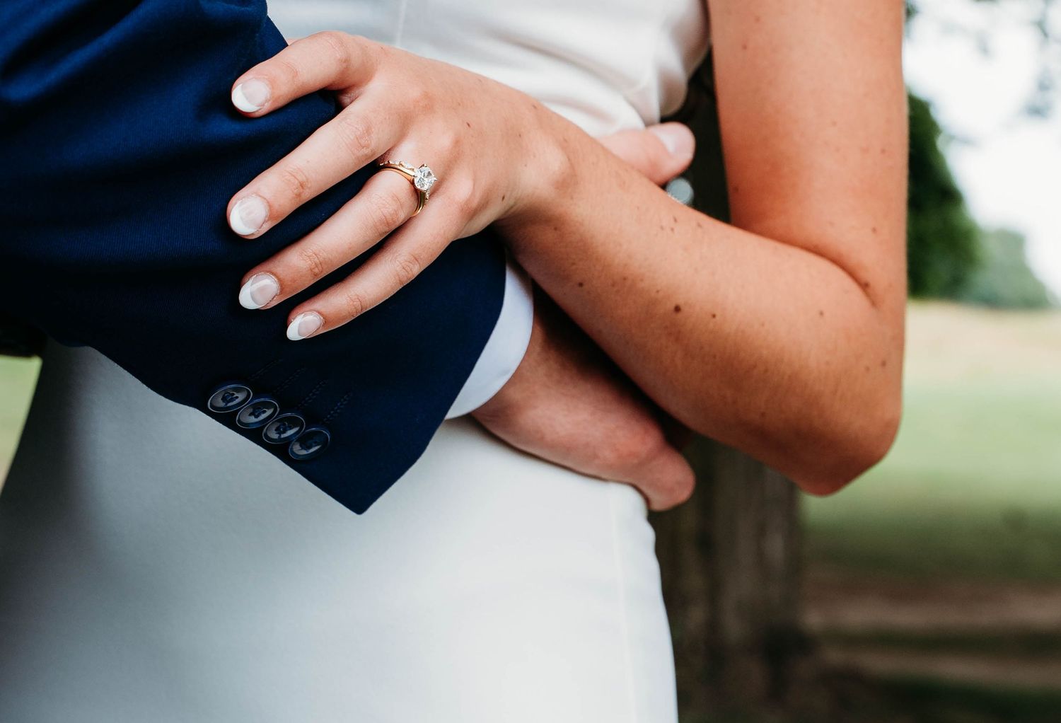 A couple holds hands during an outdoor wedding ceremony, showcasing their formal attire and wedding rings.