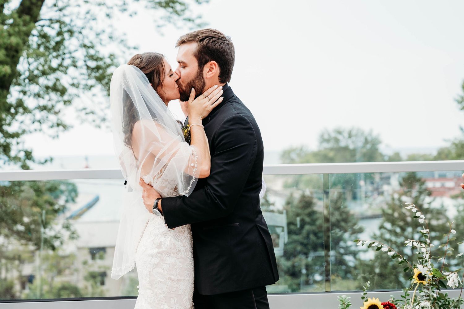 A couple in wedding attire shares a romantic kiss outdoors surrounded by lush greenery and natural light.