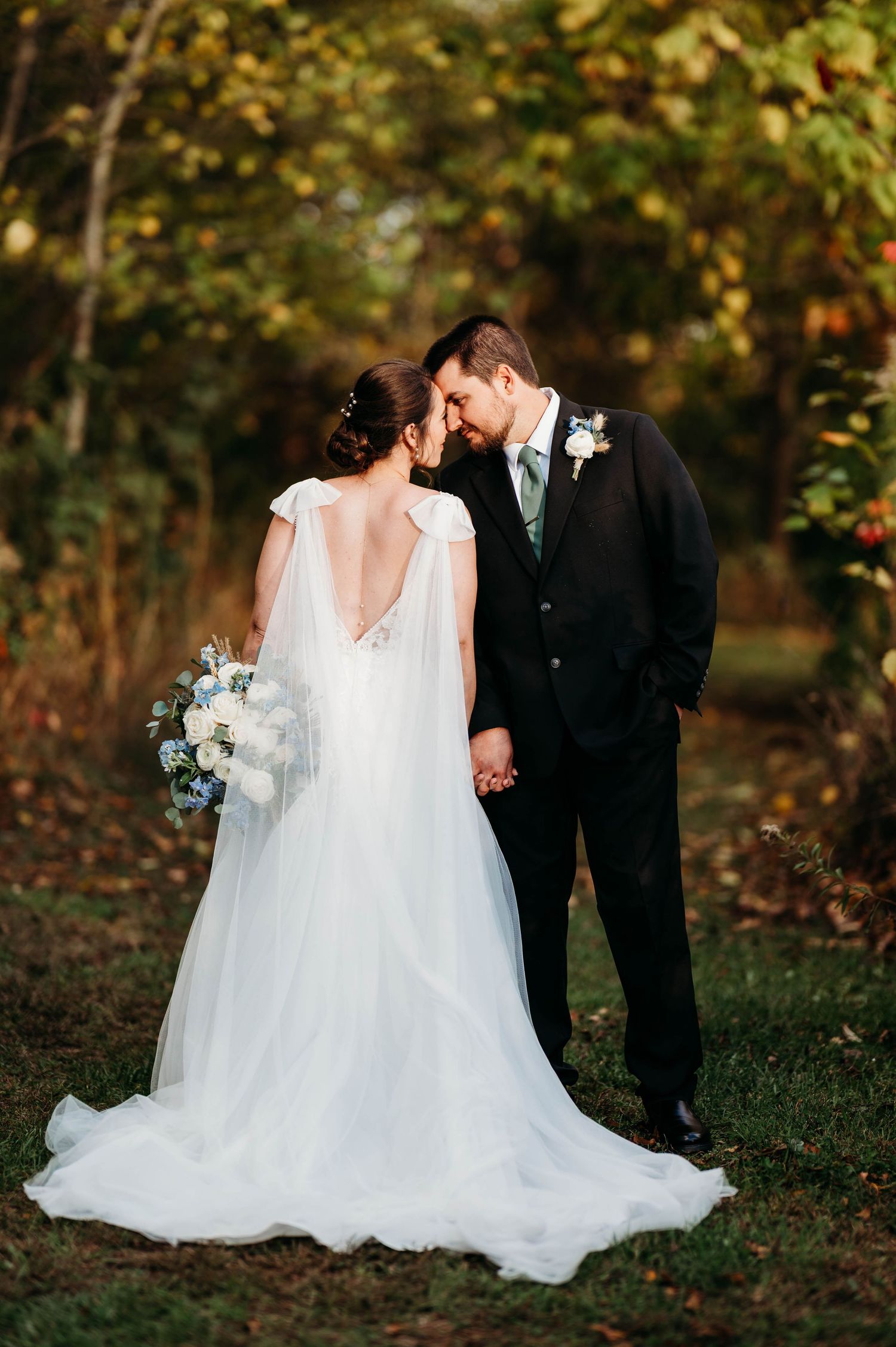 A couple shares an intimate moment in a wooded outdoor setting during their wedding, with the bride wearing a flowing white gown.