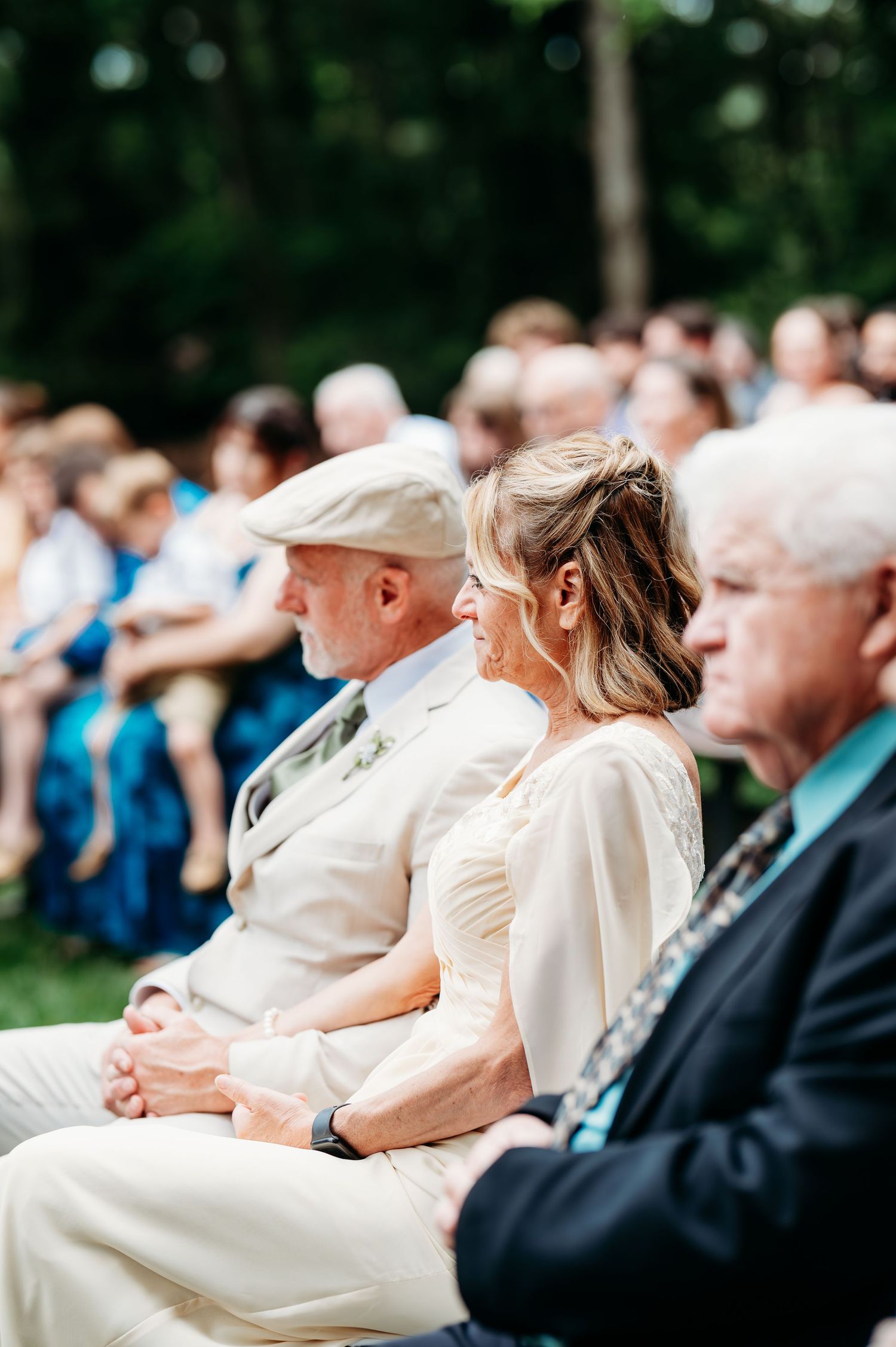 Guests seated outdoors at a wedding ceremony, smiling and watching the event unfold in a lush green setting.