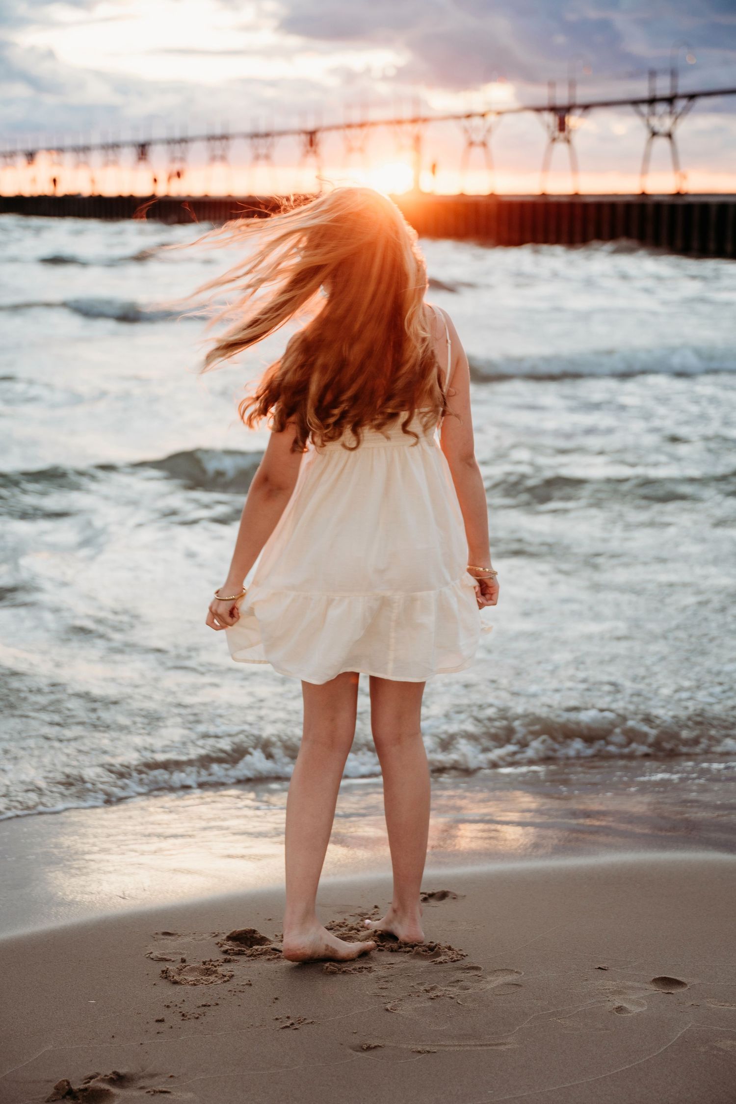 A person in a white dress walking along the sandy beach at sunset while their hair flows in the ocean breeze.