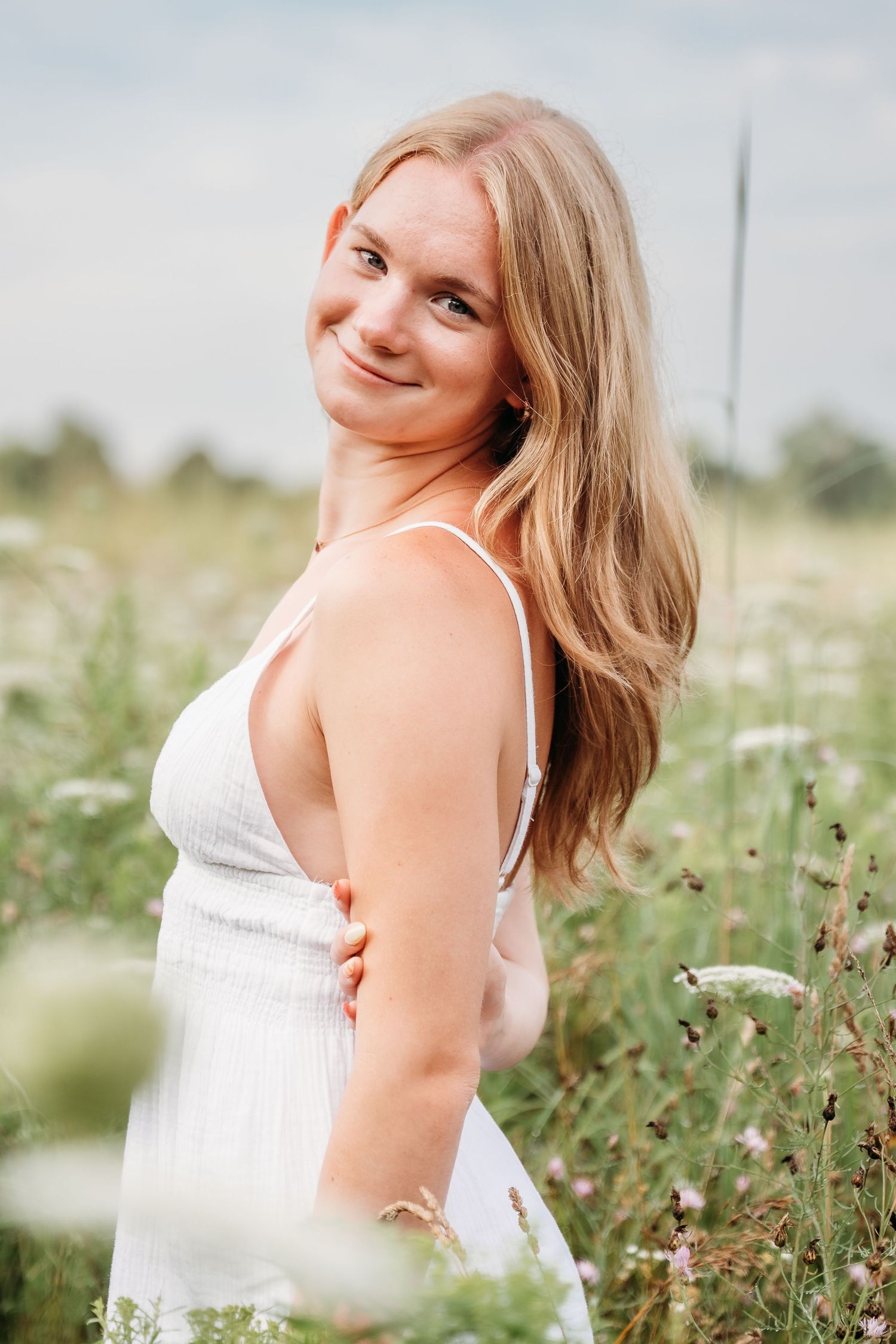 A woman in a white dress poses playfully in a wildflower meadow with soft summer lighting.