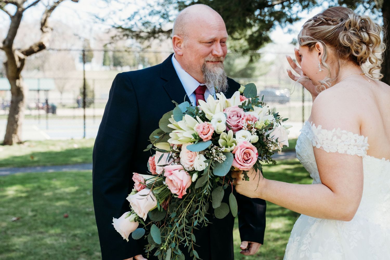 A touching moment at an outdoor wedding ceremony with a beautiful bouquet of pink roses and greenery.