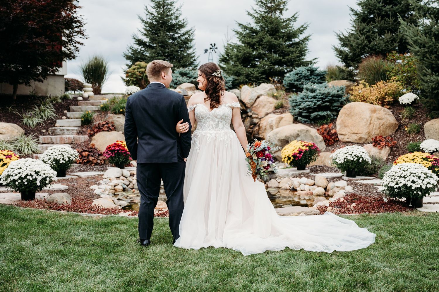 A couple in wedding attire stands together in a beautiful garden setting with evergreen trees and colorful flowers.