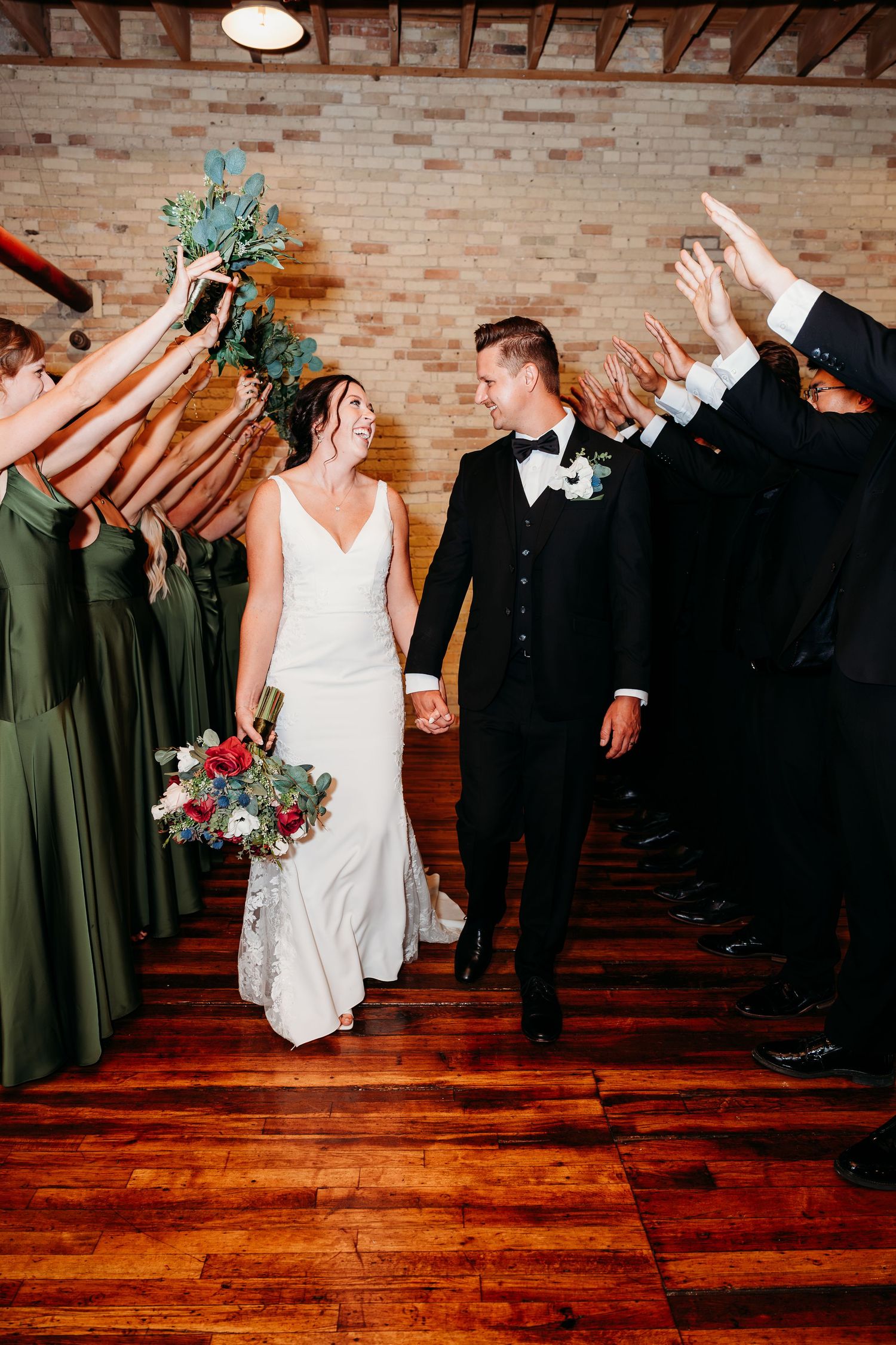 Bride and groom walk through a celebratory tunnel of hands, bride holding a lush bouquet, bridesmaids in olive green gowns.