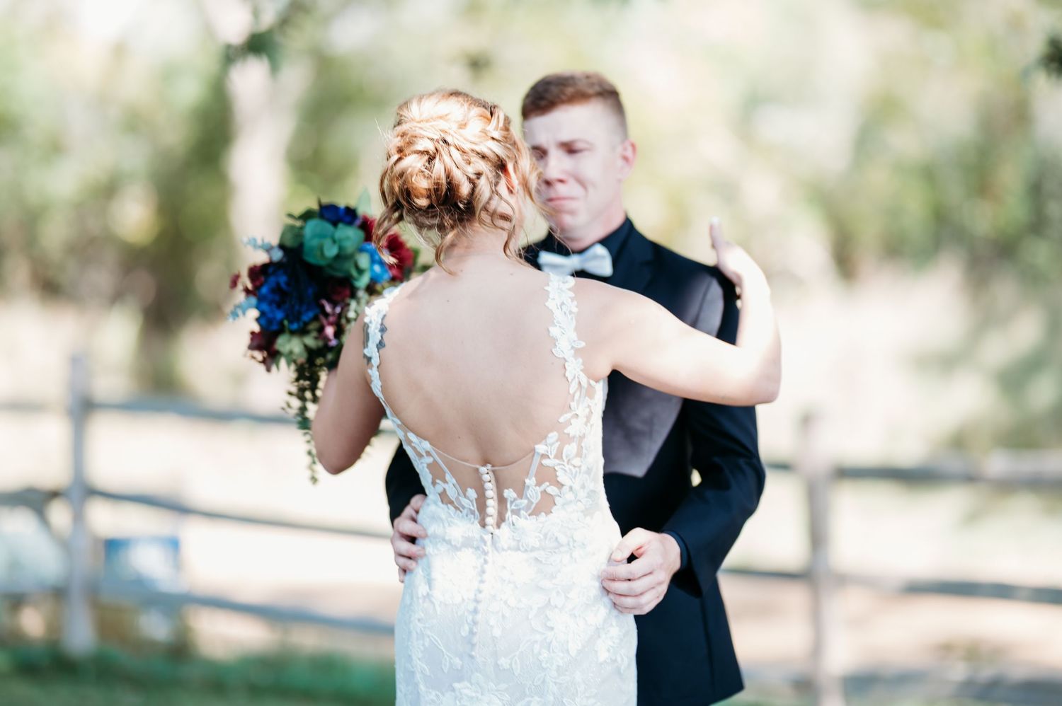 Bride and groom share first dance outdoors, bride in backless lace gown holding vibrant blue and red bouquet.