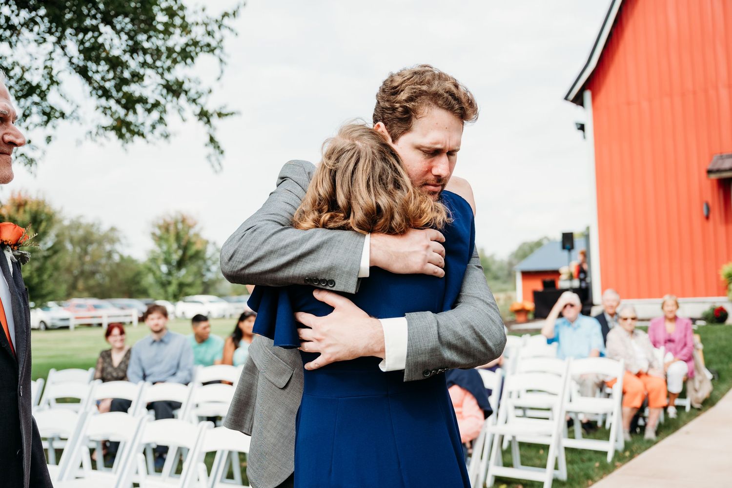 Two people share an emotional hug at an outdoor wedding ceremony with white chairs and a red barn backdrop.
