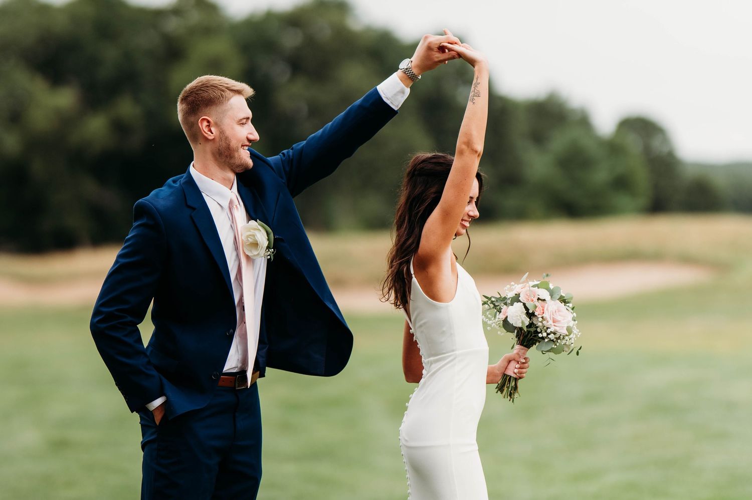 Bride and groom dancing outdoors on golf course, groom in navy suit twirling bride in white gown holding bouquet.