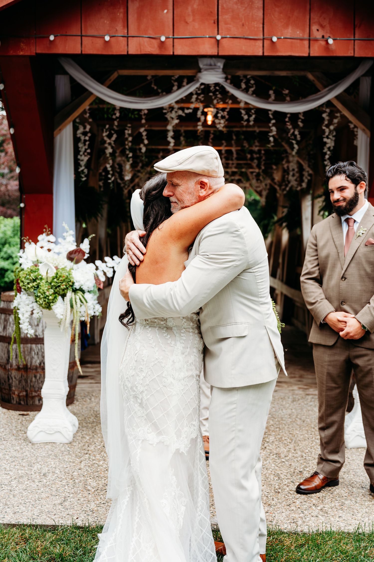 Bride and older man share emotional hug at outdoor barn wedding ceremony, with guests watching in background.