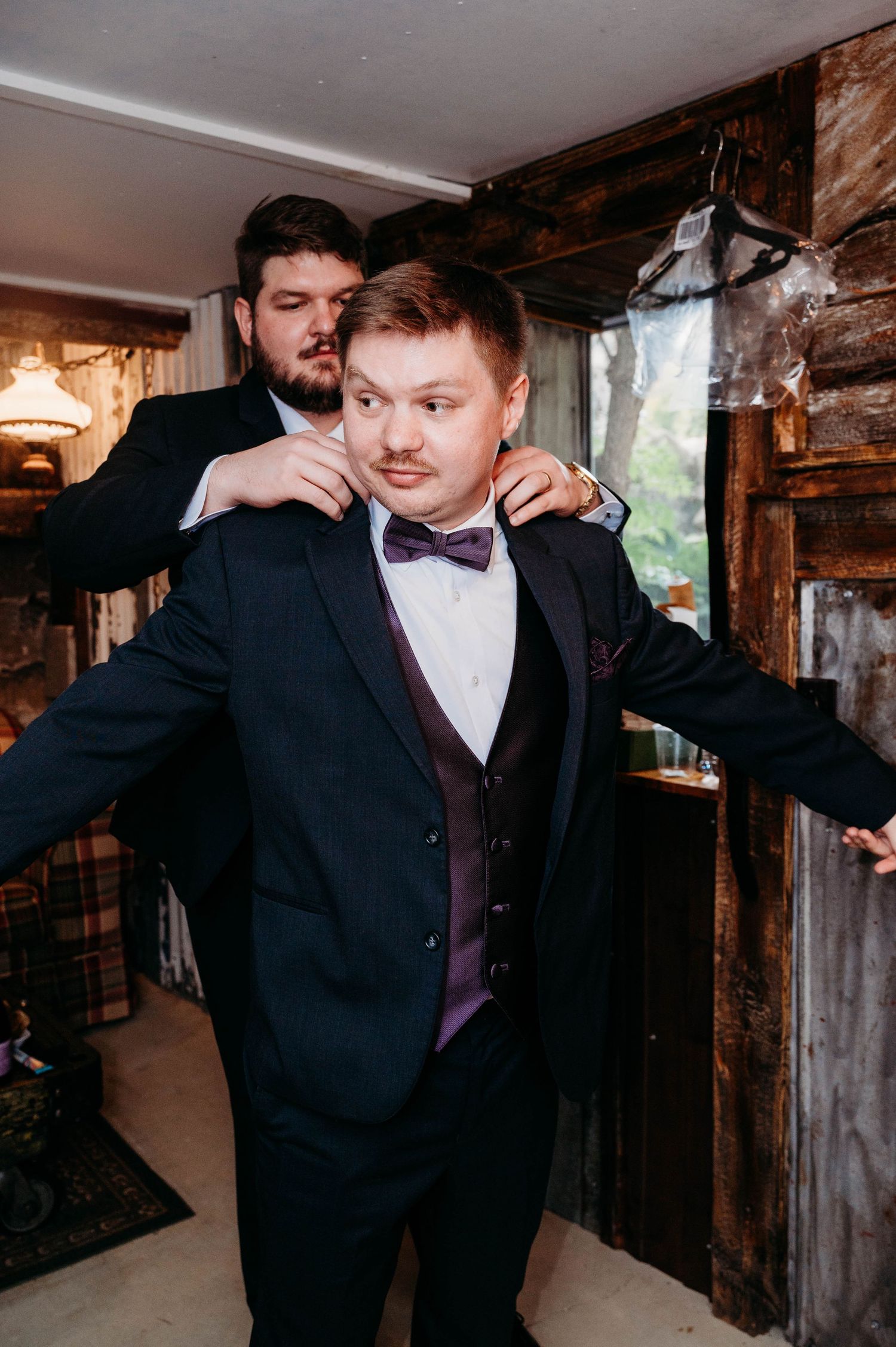 Groomsmen help adjust formal attire in rustic wooden interior before wedding ceremony.