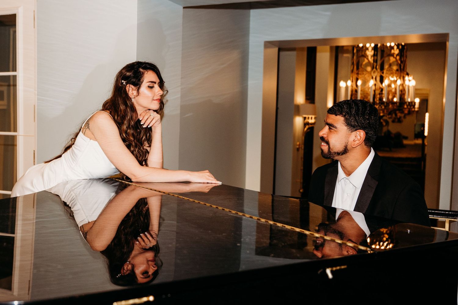 Two people share an intimate moment at a grand piano, with a chandelier glowing warmly in the background.