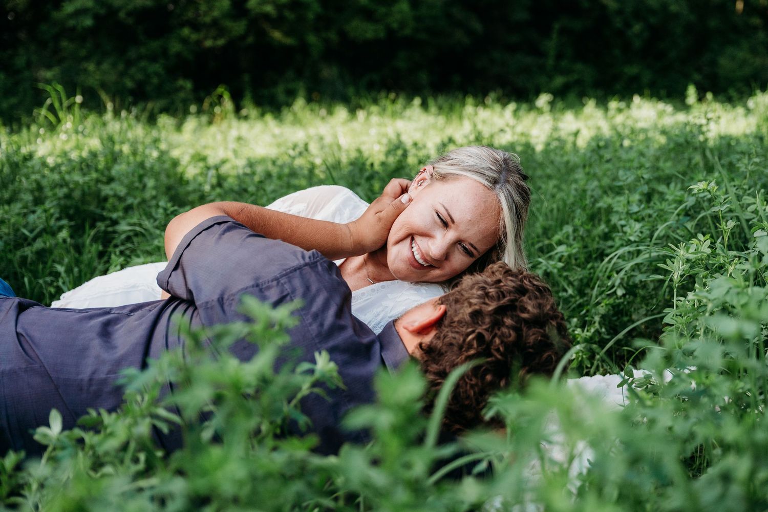 Couple laying together on grass surrounded by greenery and small purple flowers on a sunny day.