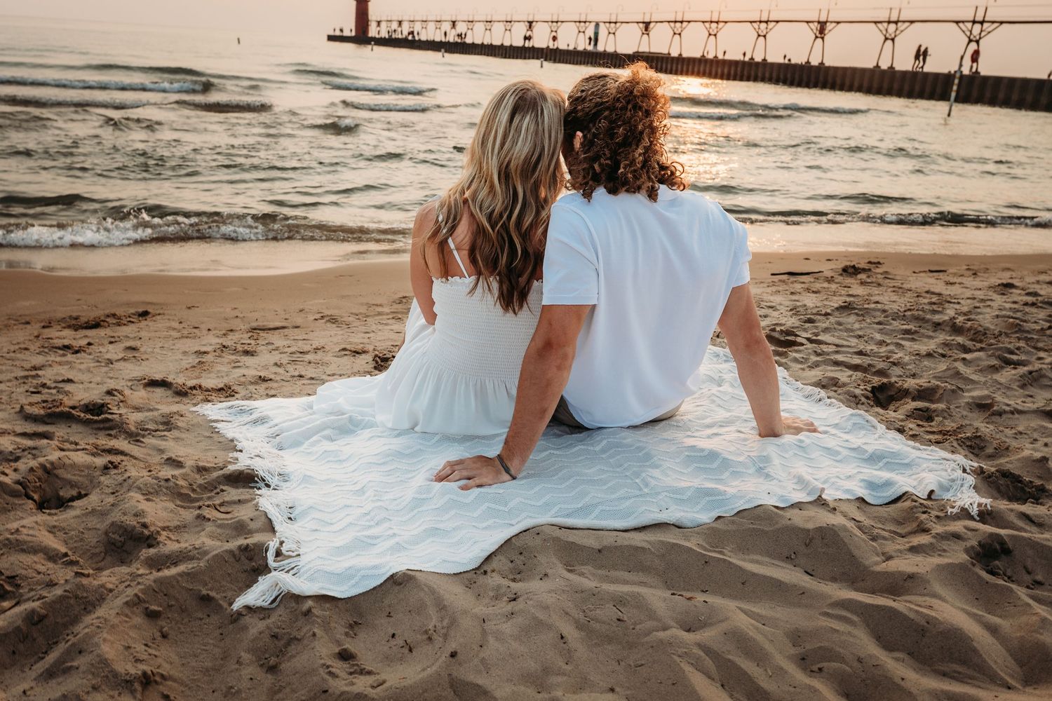 A couple sits on a white blanket at the beach during sunset with a pier in the background.