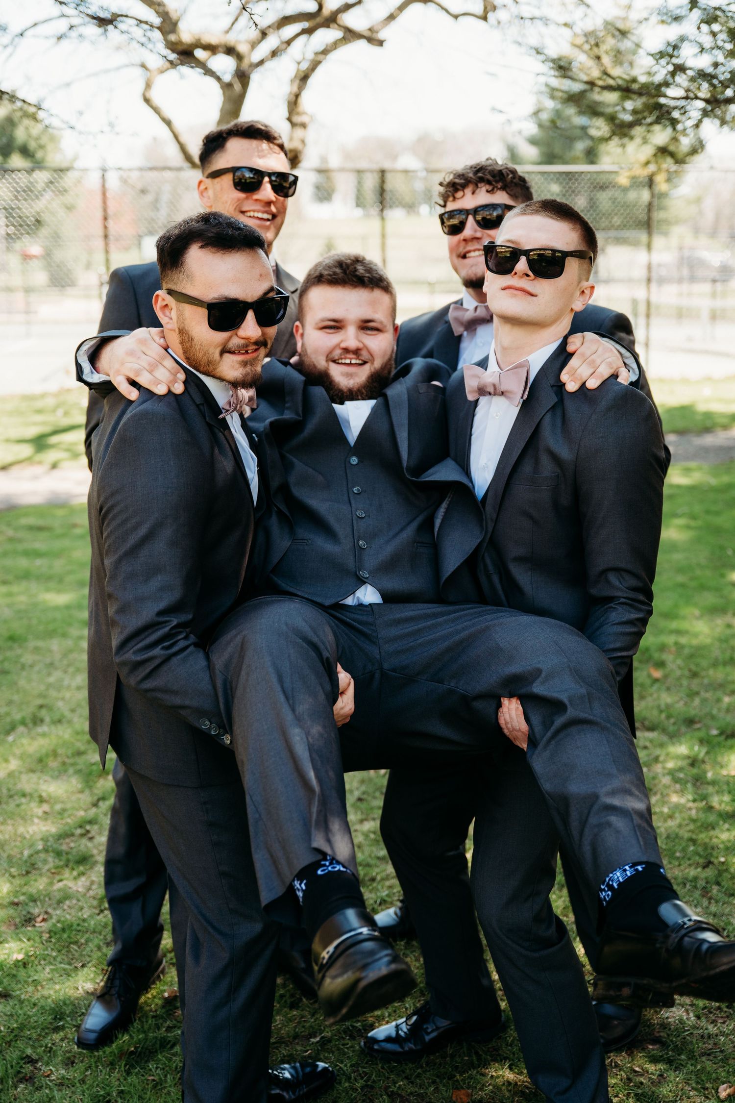 Groomsmen in black suits and sunglasses pose playfully together on grass during outdoor wedding photo session.