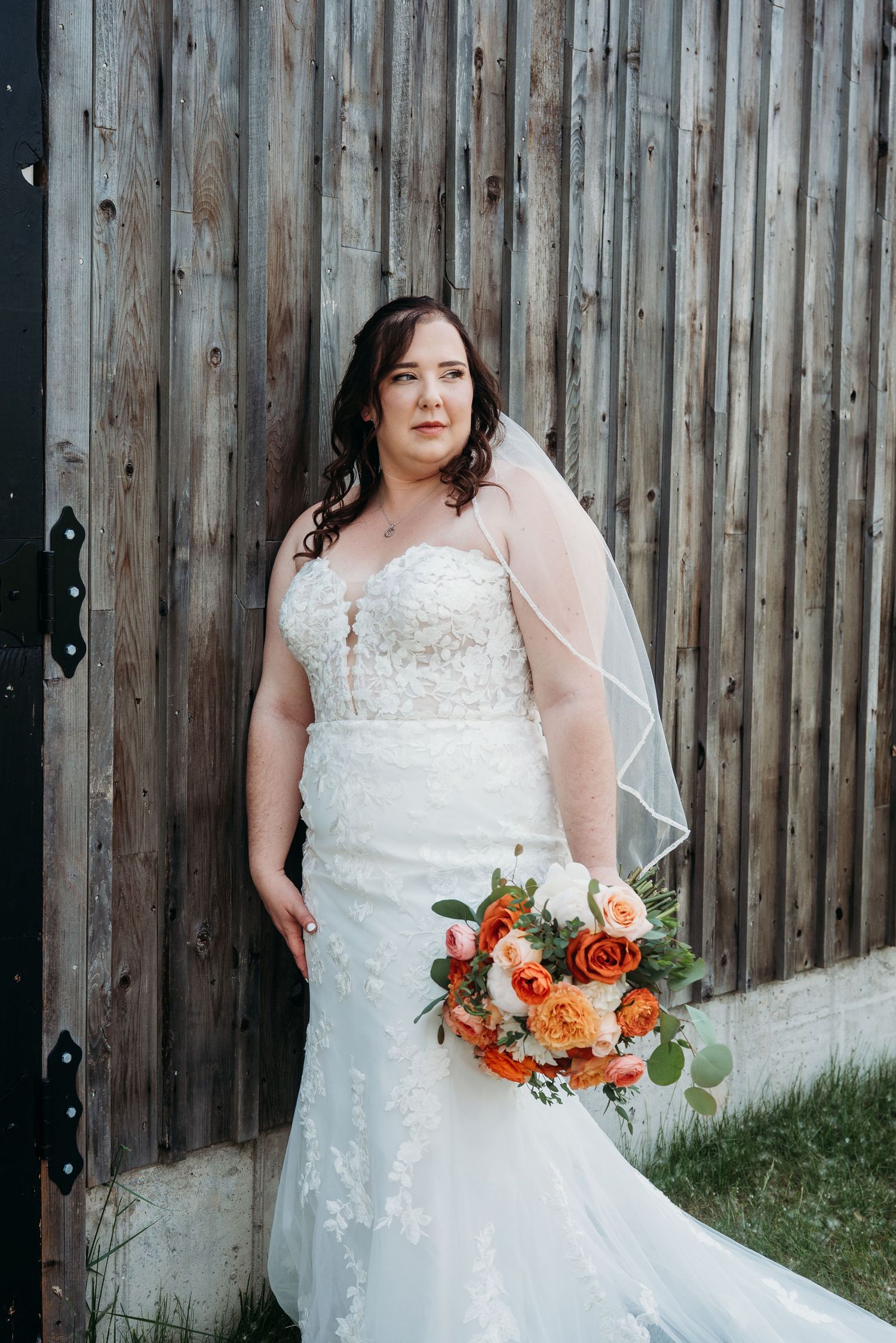 A bride in a white lace mermaid wedding dress holds a vibrant orange and red floral bouquet against a rustic wooden wall.