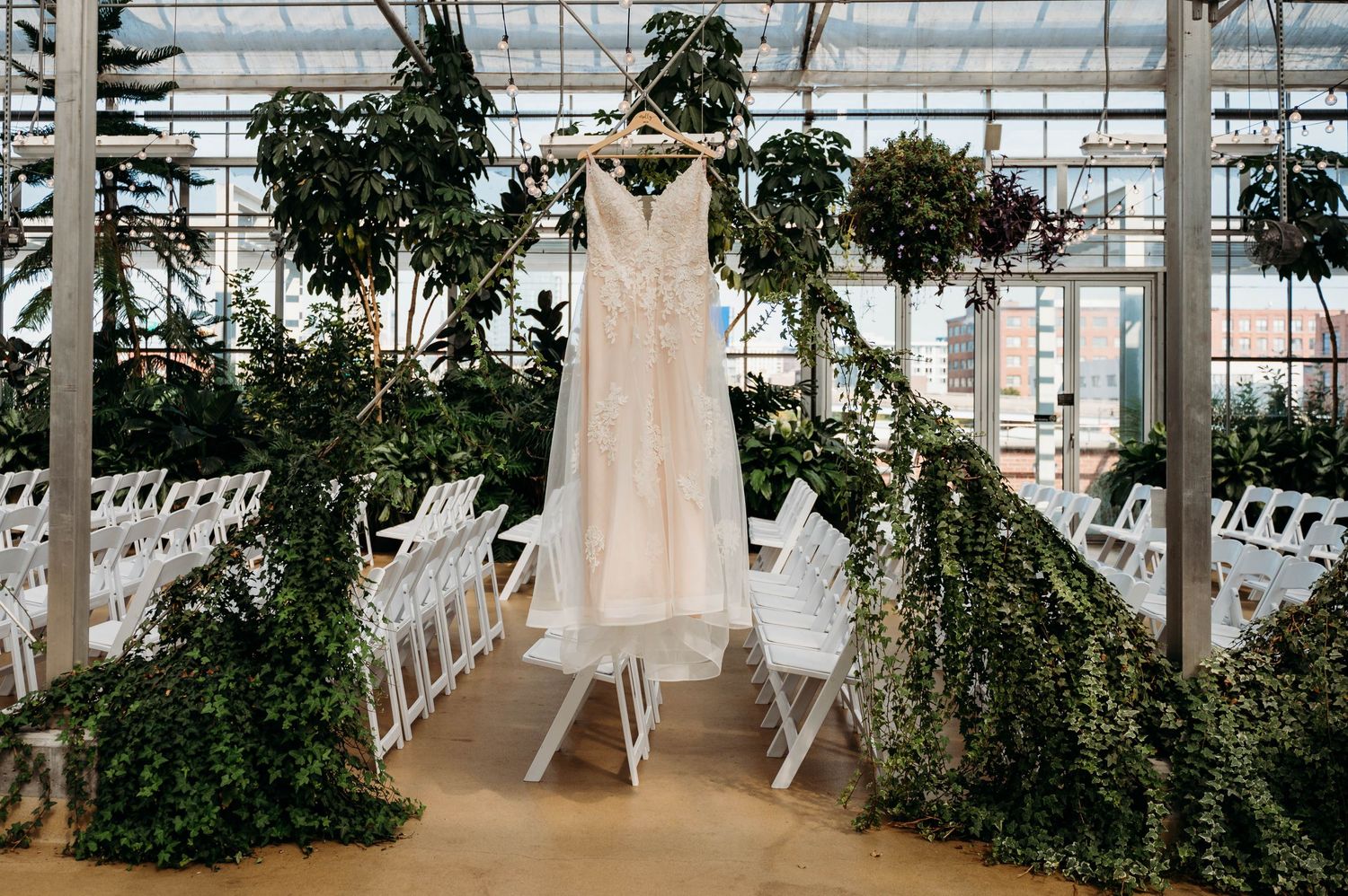 White wedding dress hangs in a greenhouse venue with white chairs and lush greenery decorations surrounding it.