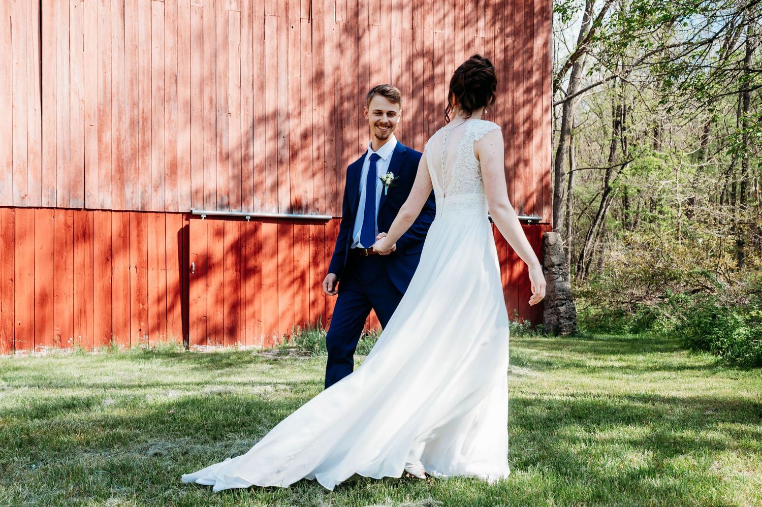 Bride in white gown holds hands with groom in navy suit outdoors near a rustic red barn on their wedding day. Wedding Lowell MI