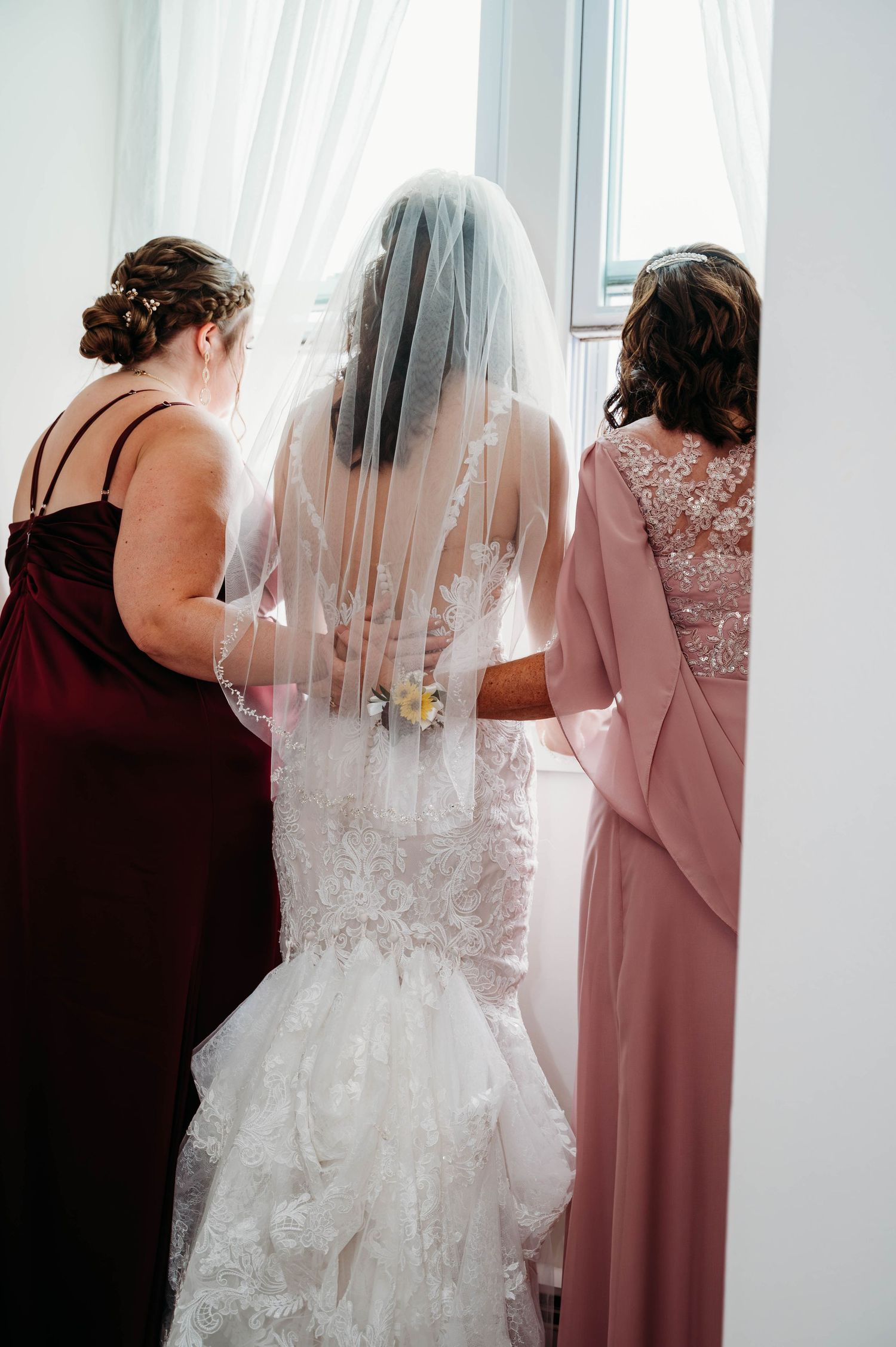 Wedding party helps bride with final dress preparations in soft pink and burgundy gowns.