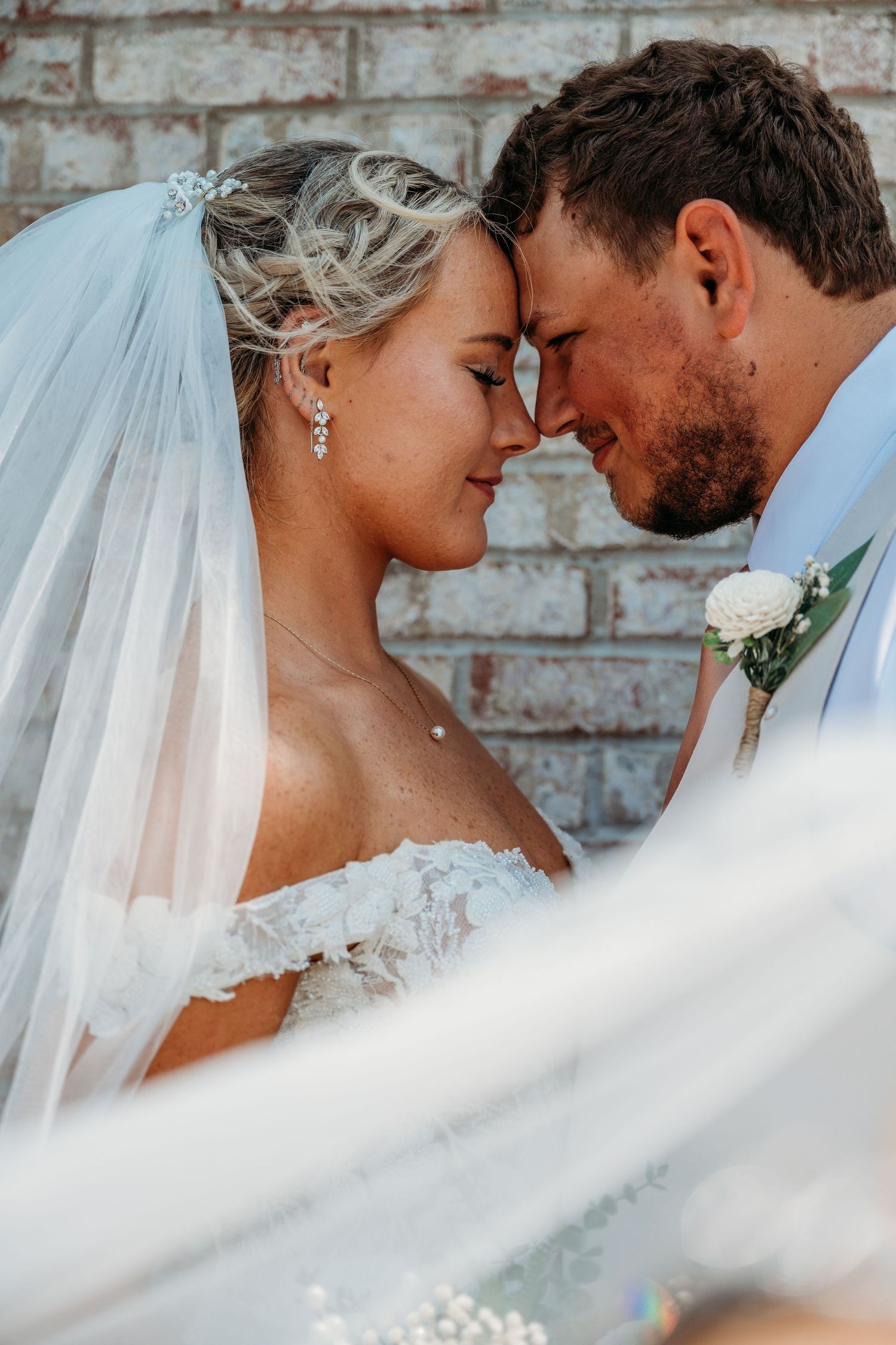 Romantic wedding portrait with flowing veil against rustic brick wall background.