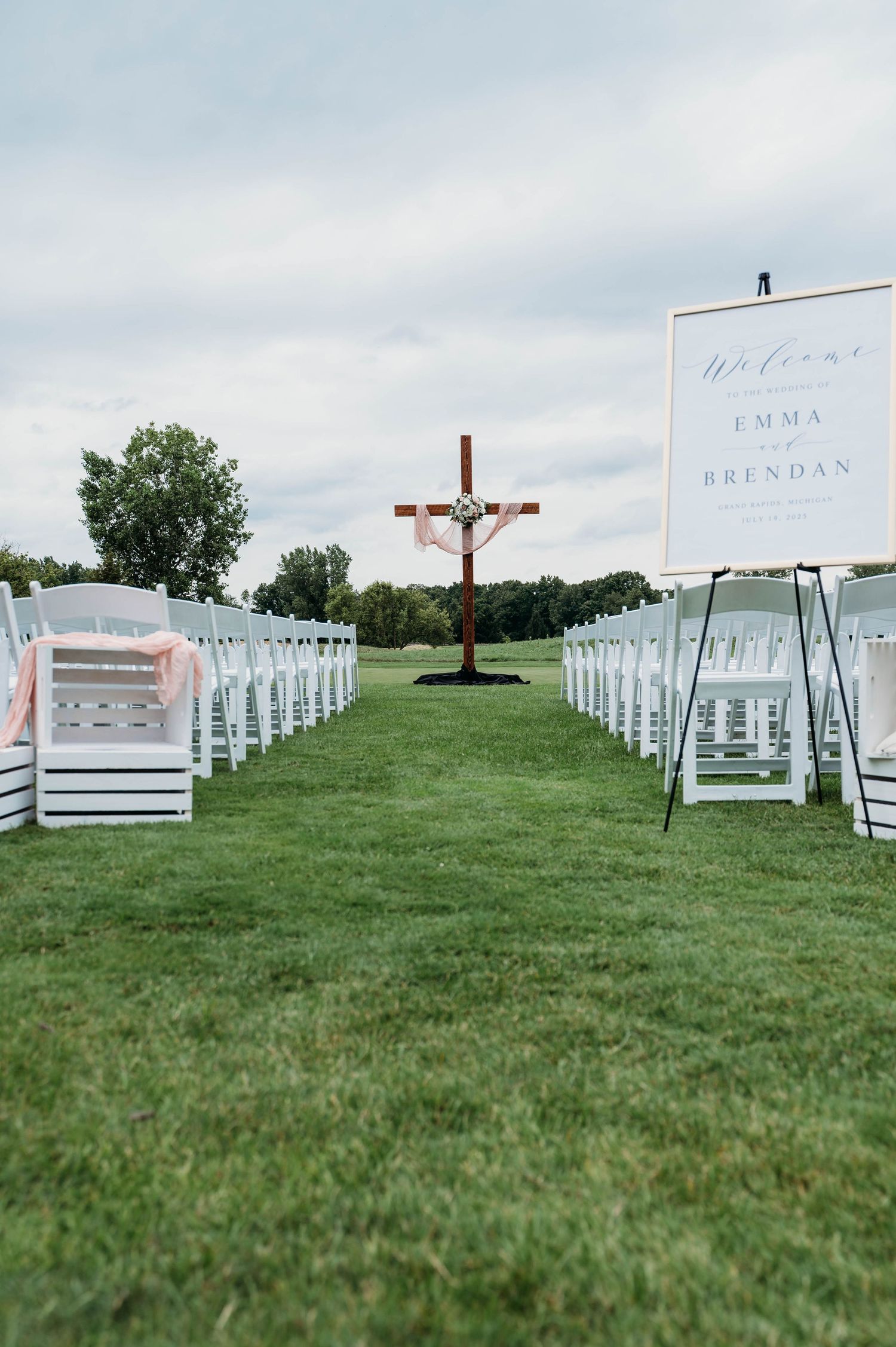 White wedding chairs arranged on a green hillside lawn with welcome signs and decorative elements for an outdoor ceremony.
