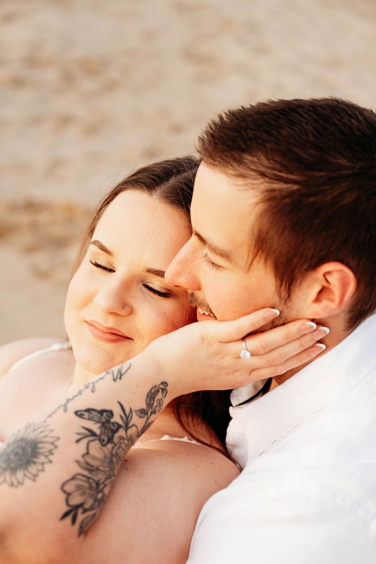 Couple embracing on beach, woman with floral arm tattoo and engagement ring, man kissing her cheek at sunset.