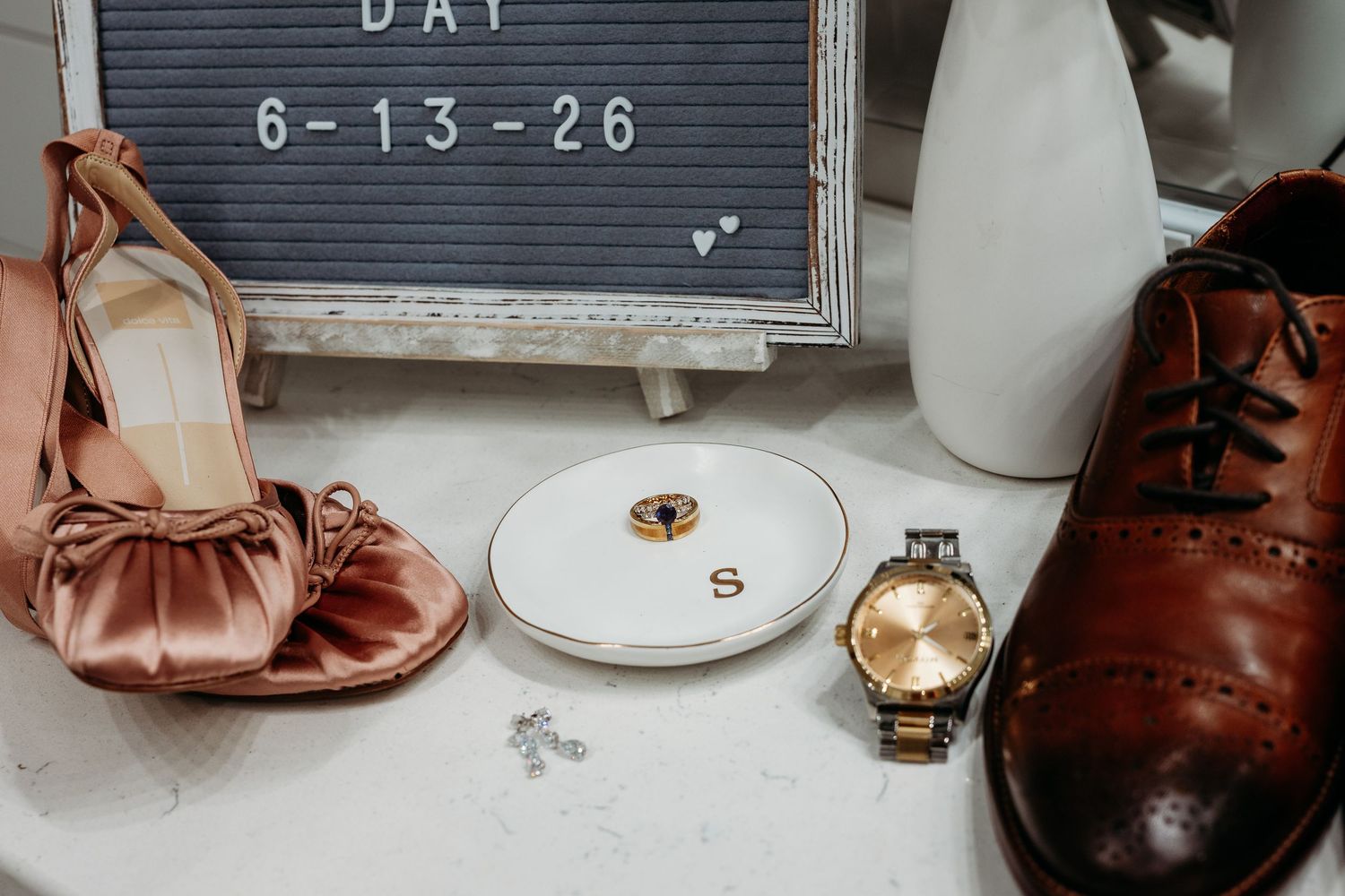 Wedding flat lay with pink satin heels, brown dress shoes, gold watch, rings on dish, and letter board reading Day 6-13-26.