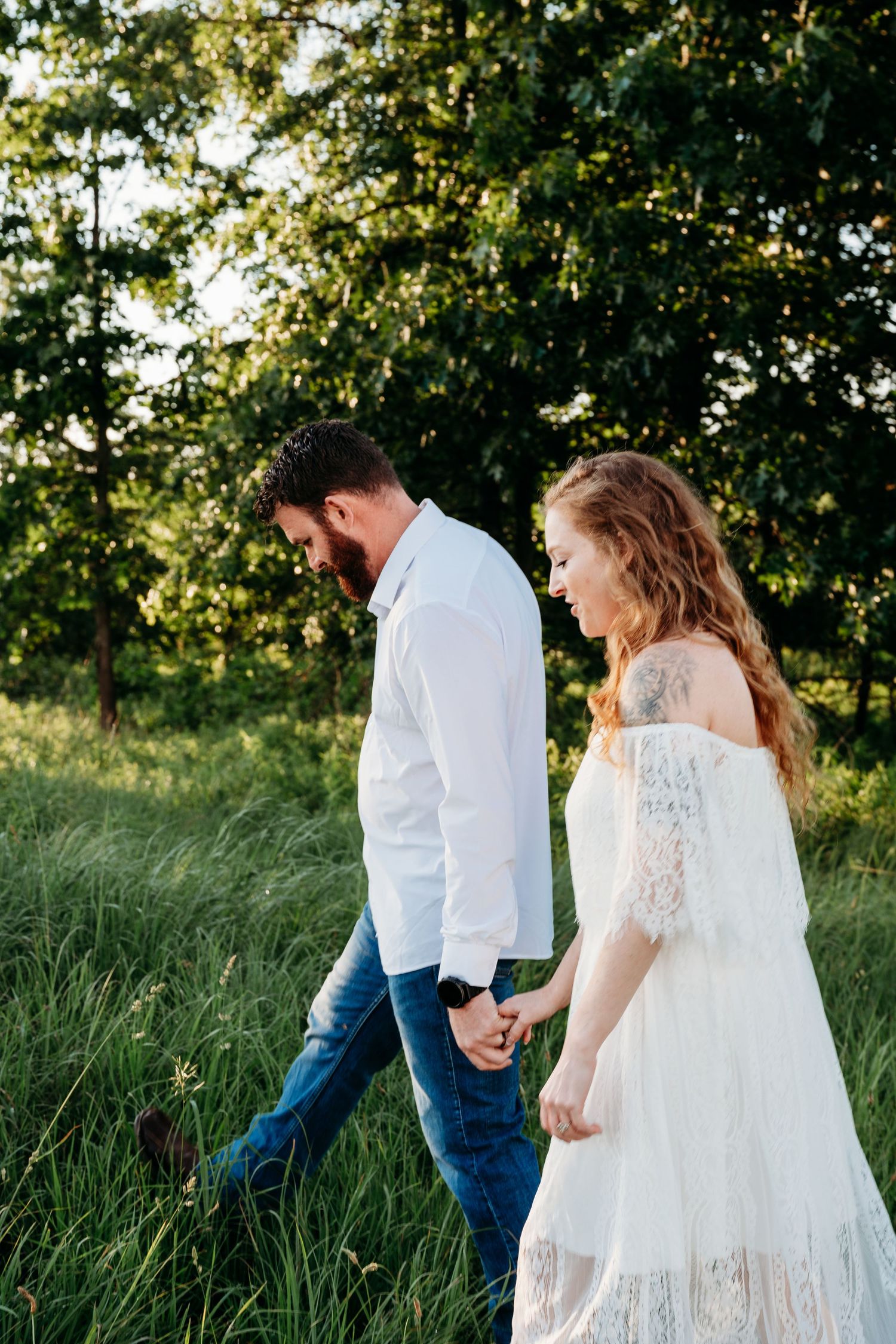 A couple in white outfits holds hands walking through a lush green field, capturing a romantic outdoor moment.