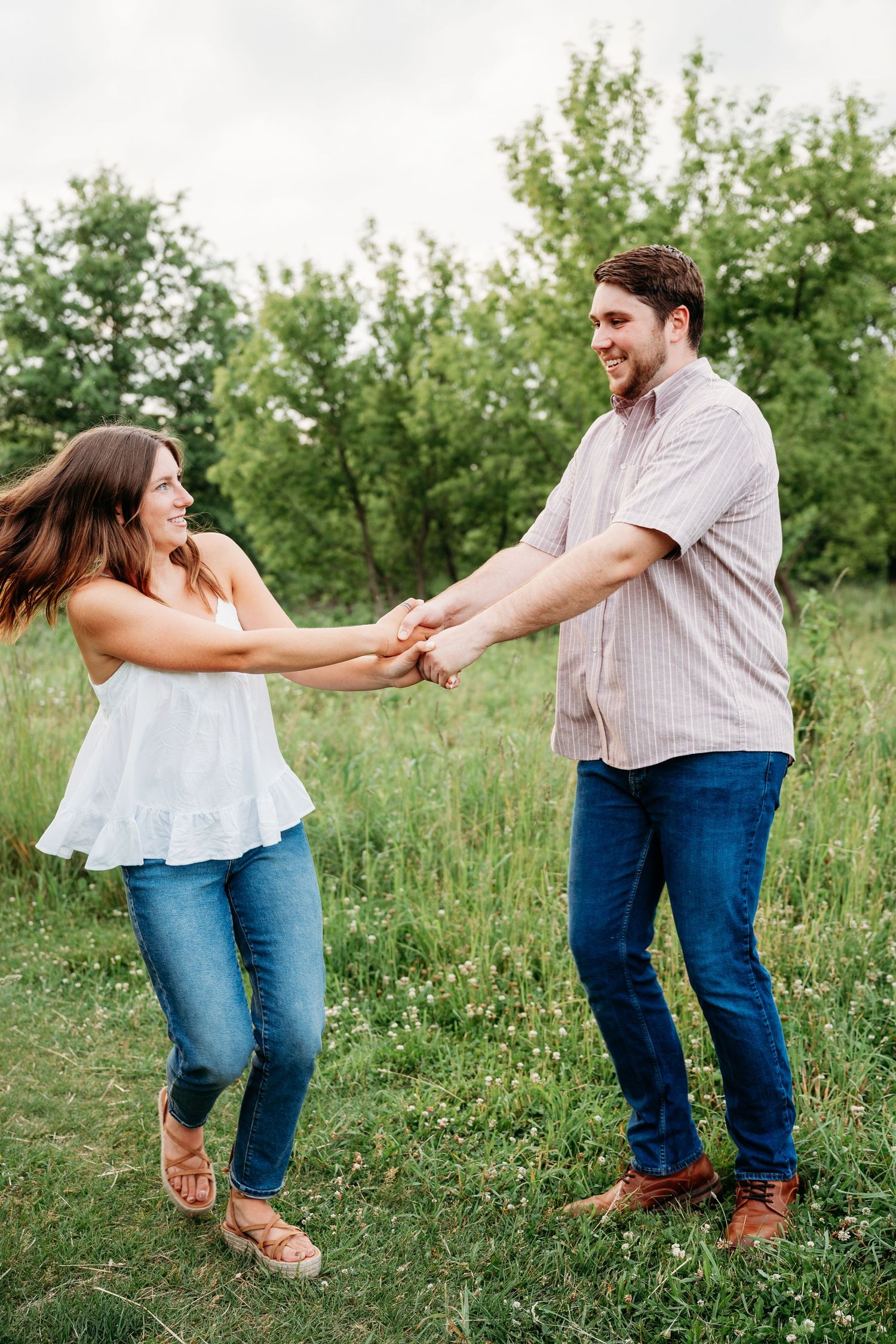 Happy couple holding hands and spinning together in a lush green field, wearing casual jeans and smiling joyfully.