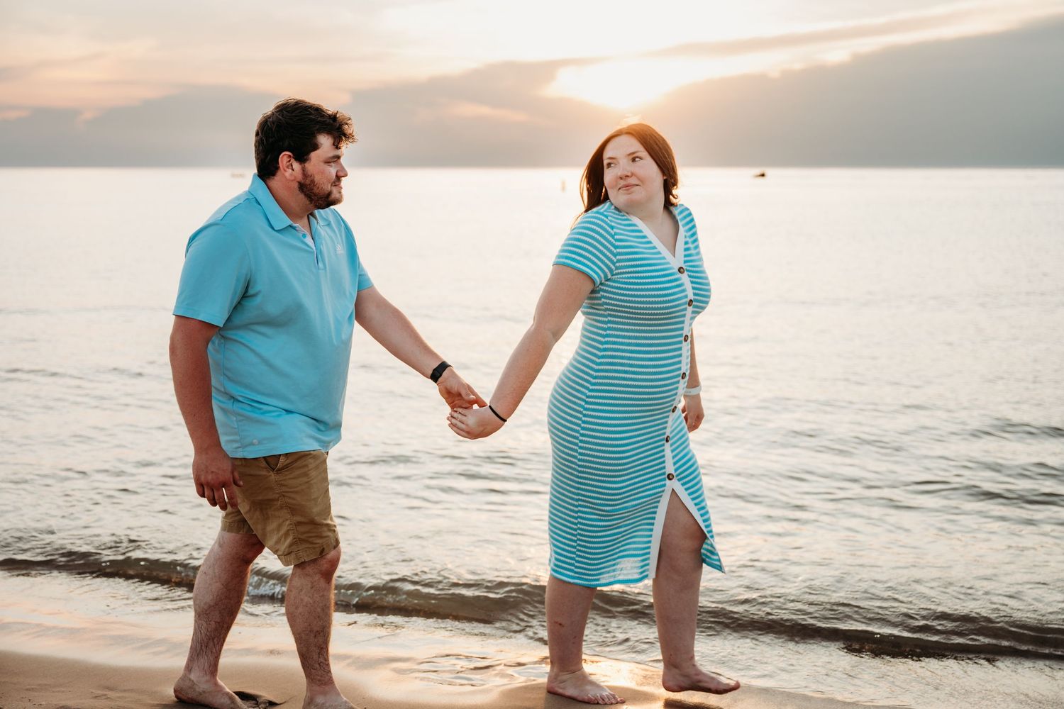 A couple walks on the beach at sunset holding hands in matching turquoise colored outfits.
