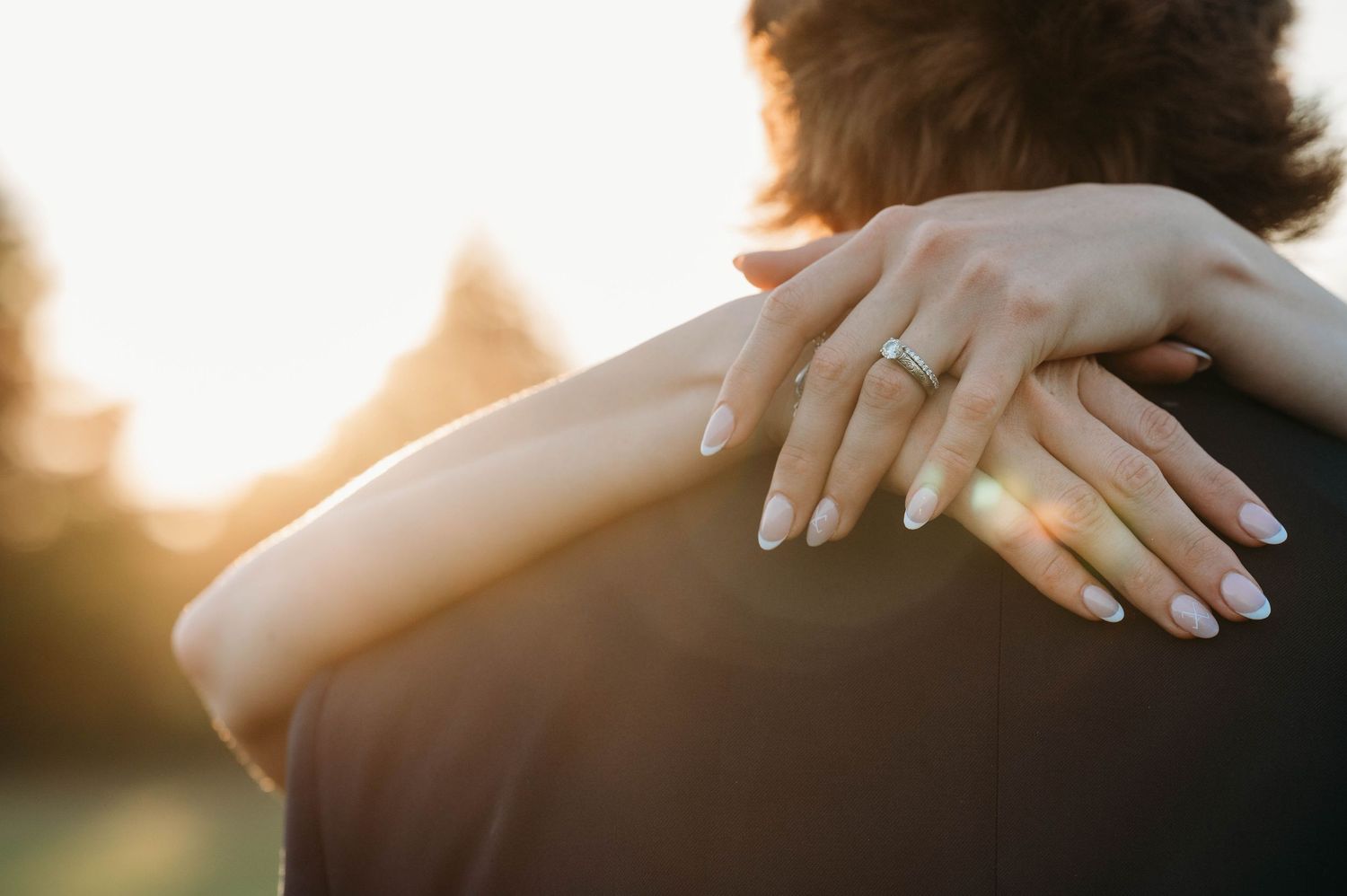 Close-up of a woman's hand with an engagement ring resting on a man's shoulder at sunset.