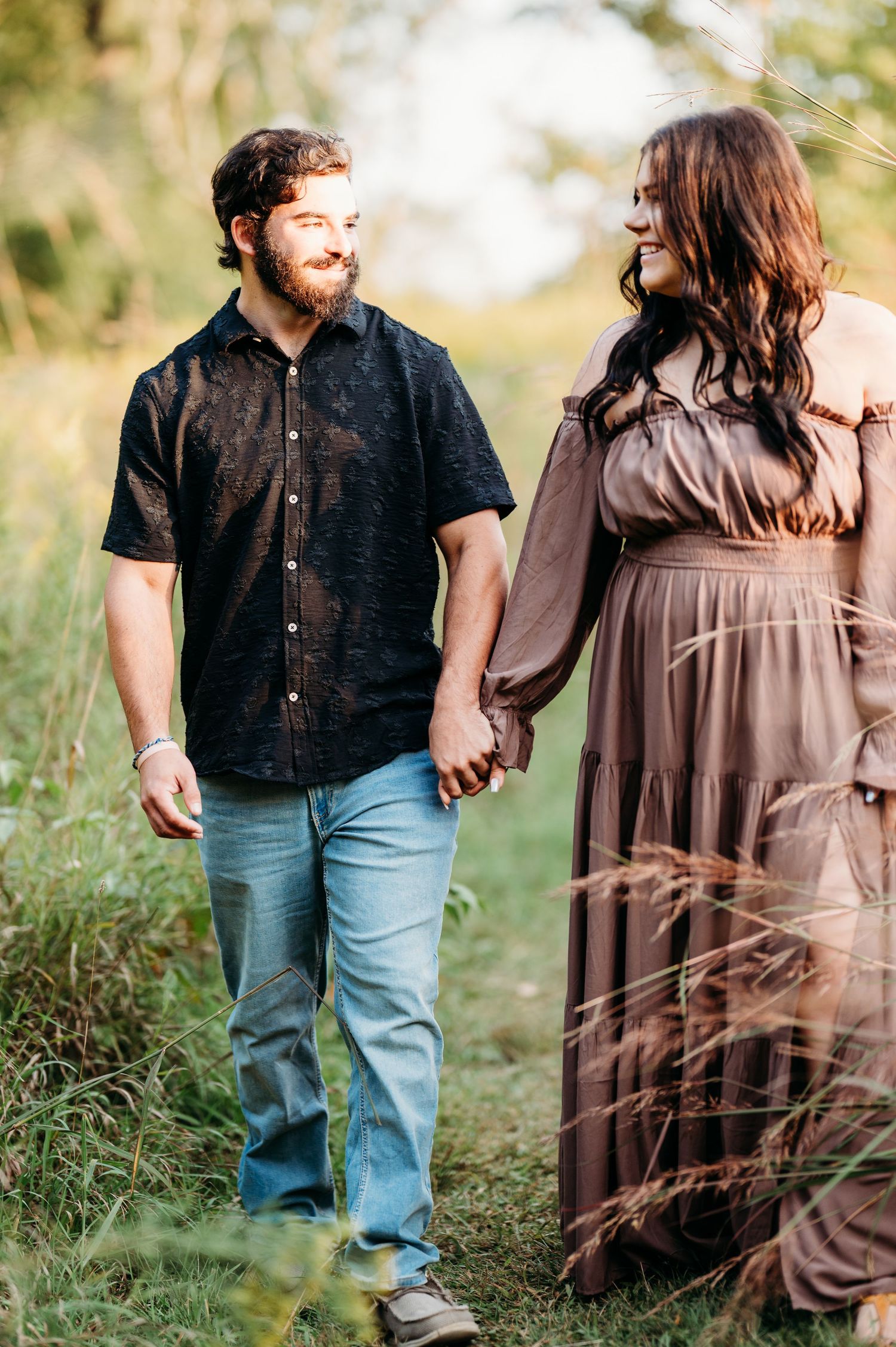 A couple holds hands and smiles at each other while walking through a lush green field during golden hour.