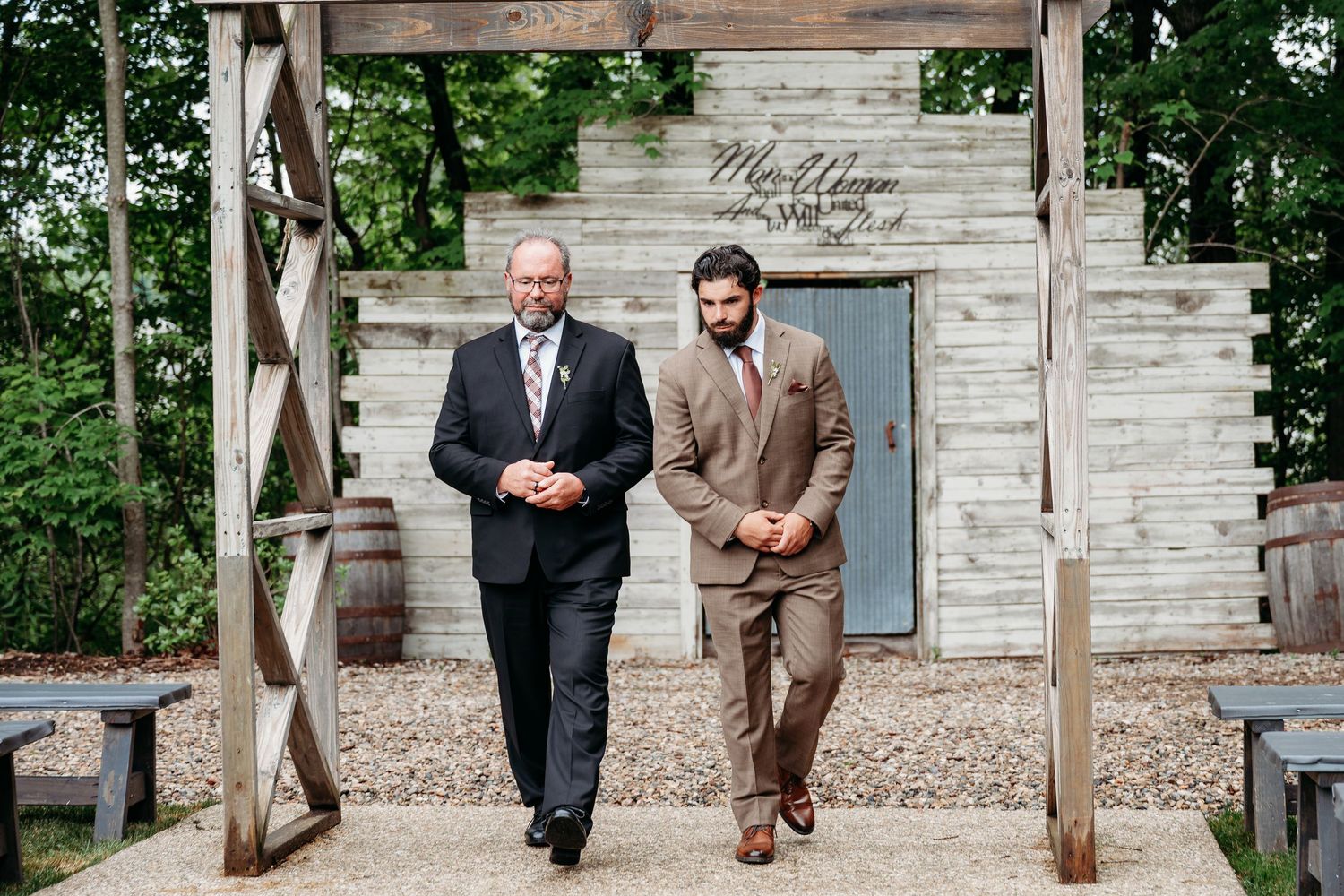 Two men in suits walk together at a rustic outdoor wedding ceremony venue with wooden decor.