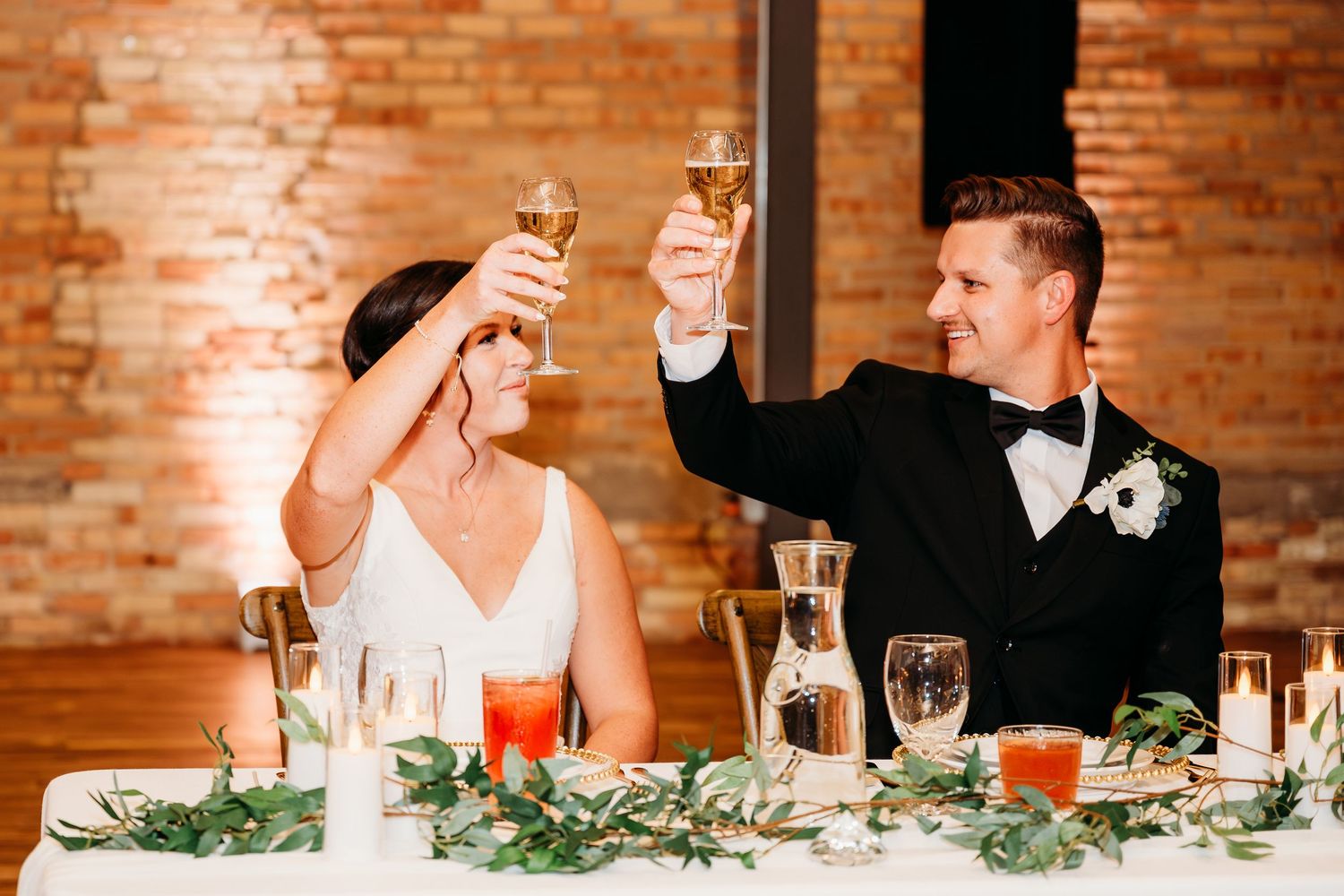 Bride and groom raising champagne glasses in a toast at their wedding reception with brick wall backdrop.