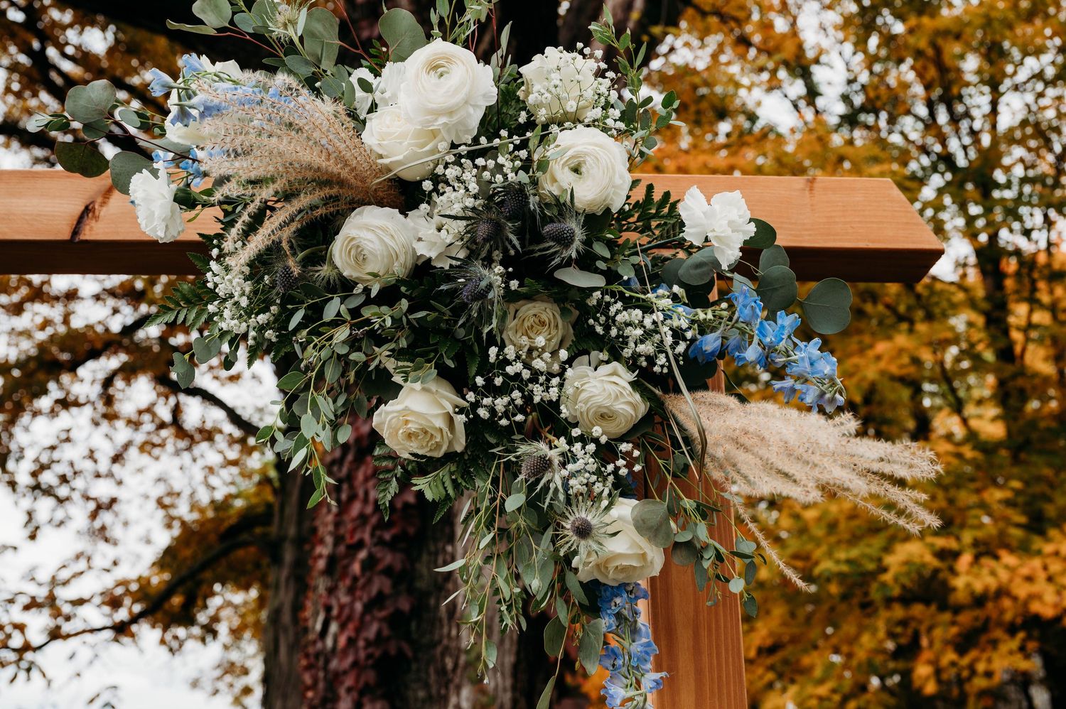 Rustic wedding arch adorned with white roses, baby's breath, eucalyptus, and pampas grass against autumn foliage backdrop.