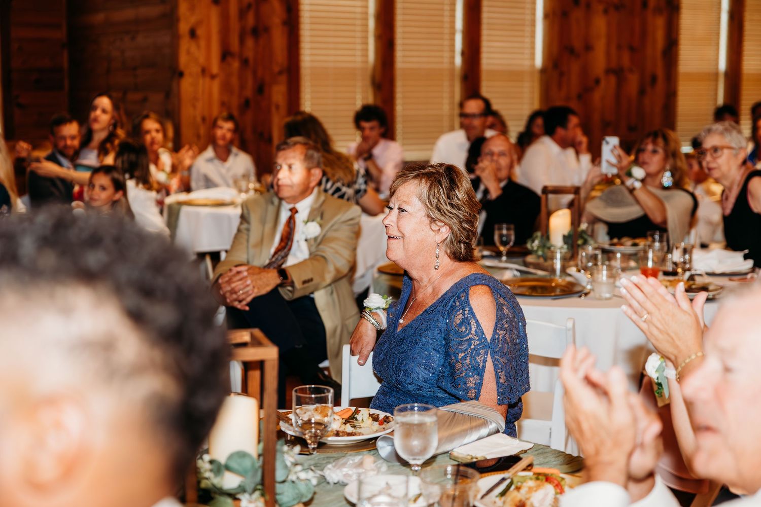 Wedding reception guests seated at decorated tables, woman in blue lace dress speaking with microphone at elegant barn venue.