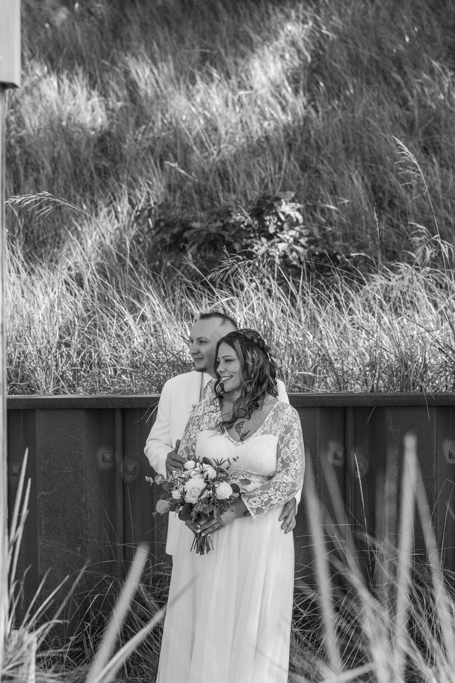 Black and white photo of a bride and groom embracing outdoors, bride holding a bouquet, with tall grass in the background.