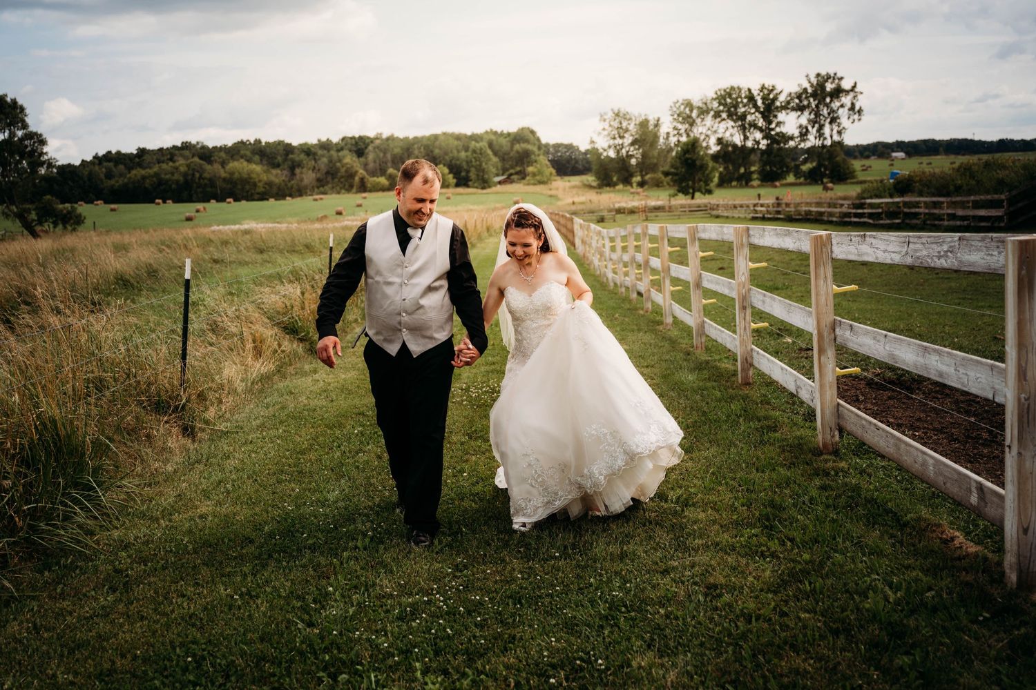 A couple walks hand in hand along a grassy path beside a white wooden fence on their wedding day.