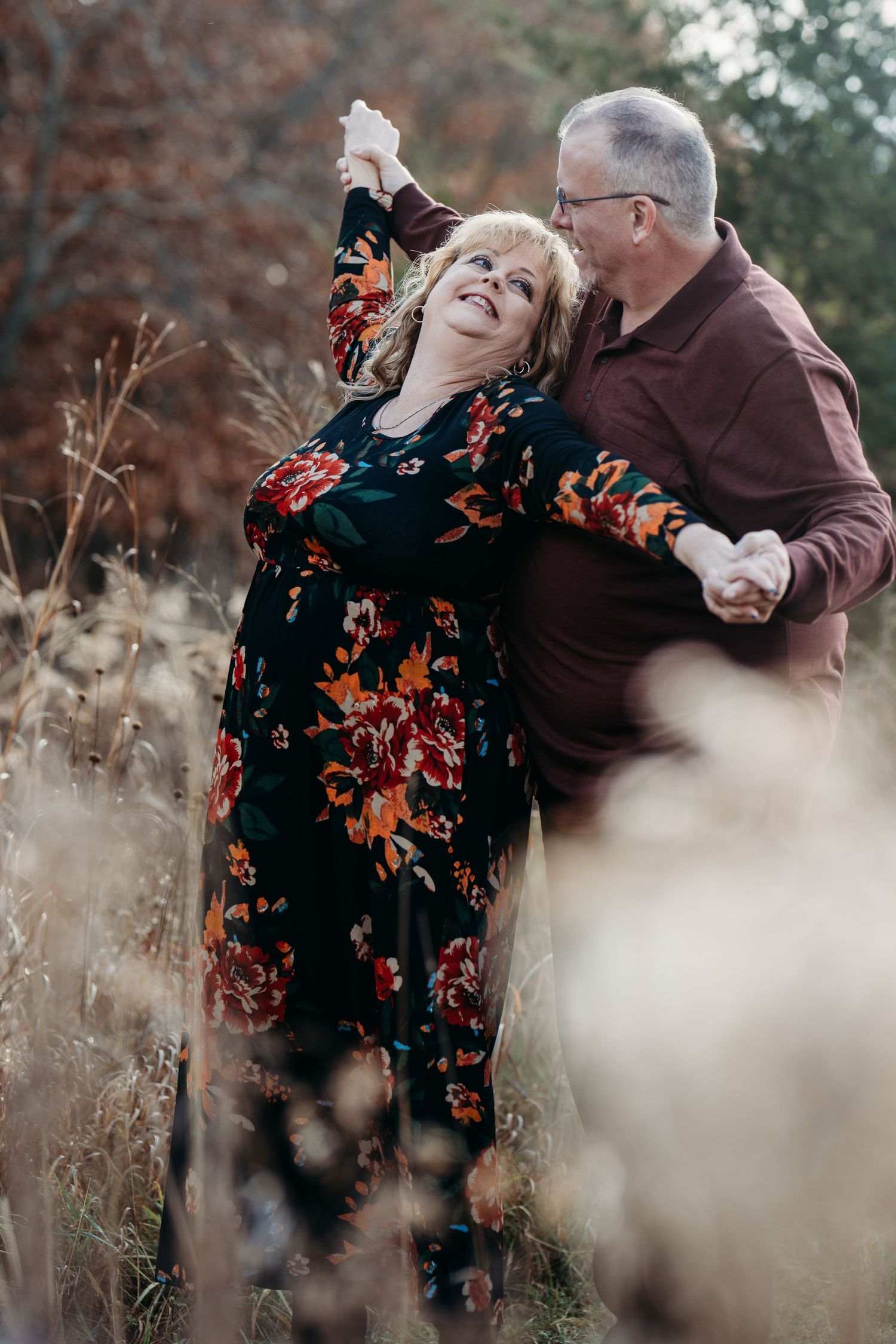 Romantic mature couple embraces in tall autumn grass as woman in floral dress leans back joyfully.