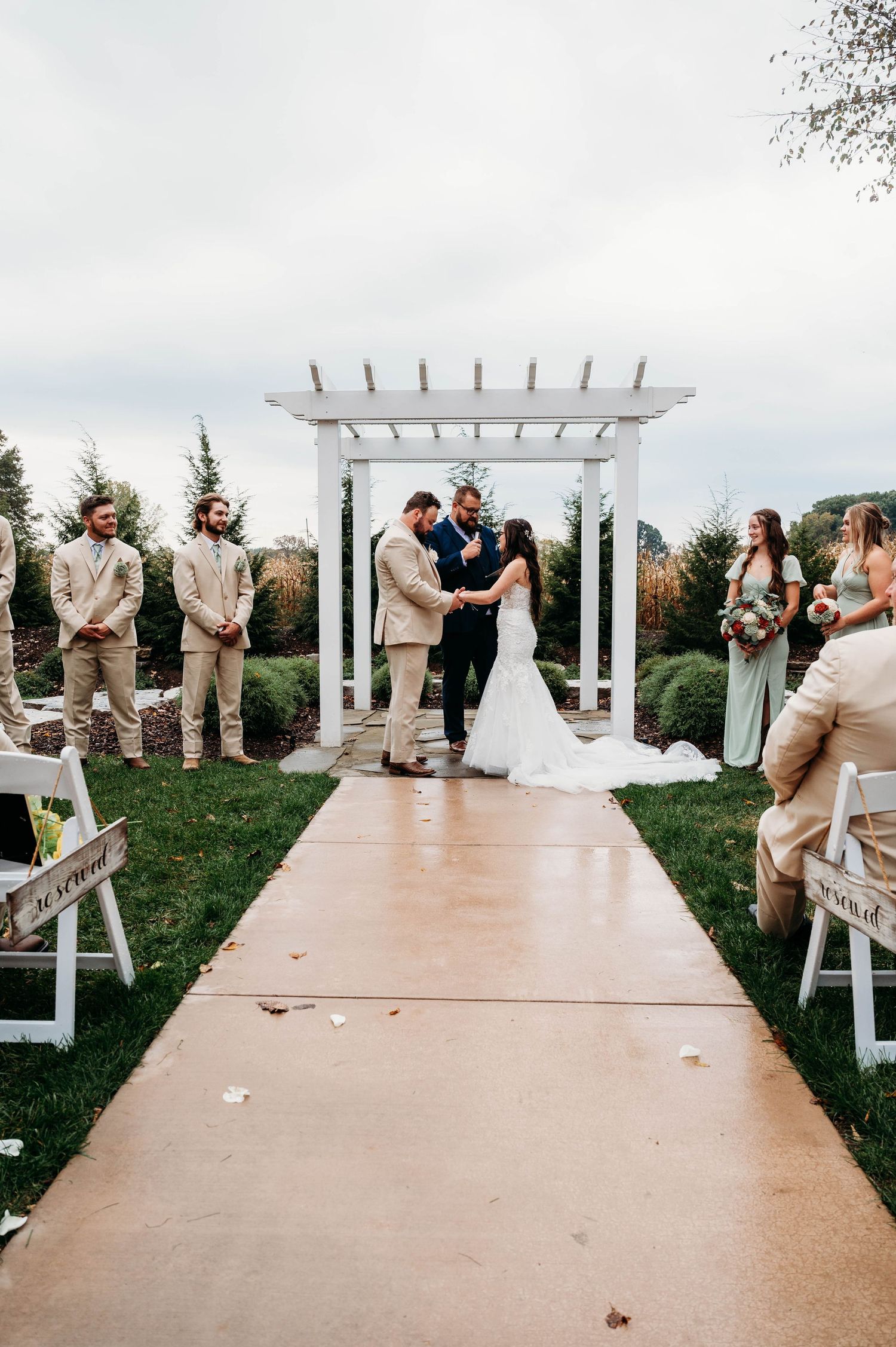 Wedding ceremony under a white pergola with guests seated on white chairs along a paved aisle.