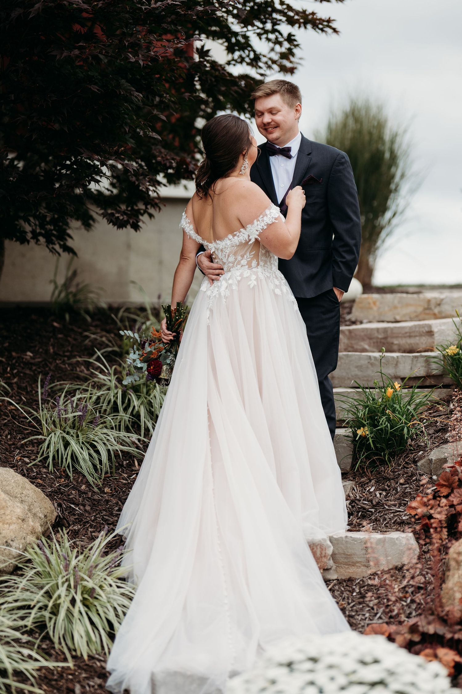 A bride and groom share an intimate moment outdoors in their wedding attire among garden landscaping and stone steps.