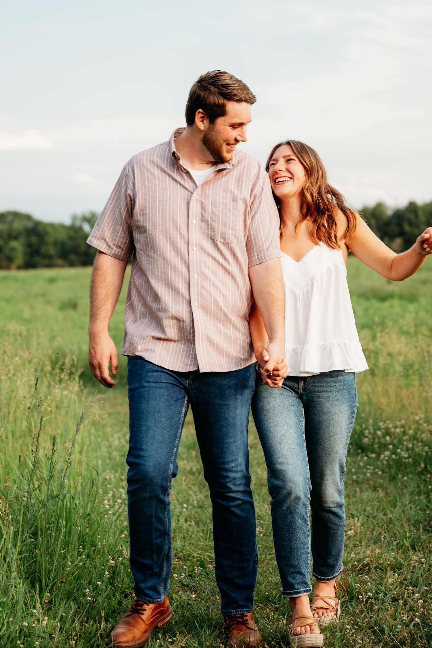 A smiling couple holds hands while walking through a lush green field on a sunny day.