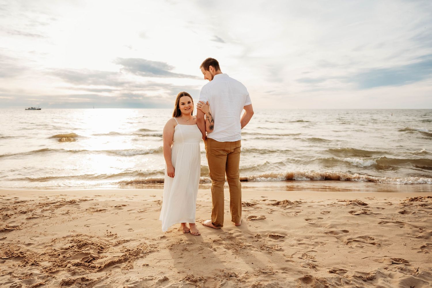Couple standing on sandy beach at sunset, facing the ocean waves together.