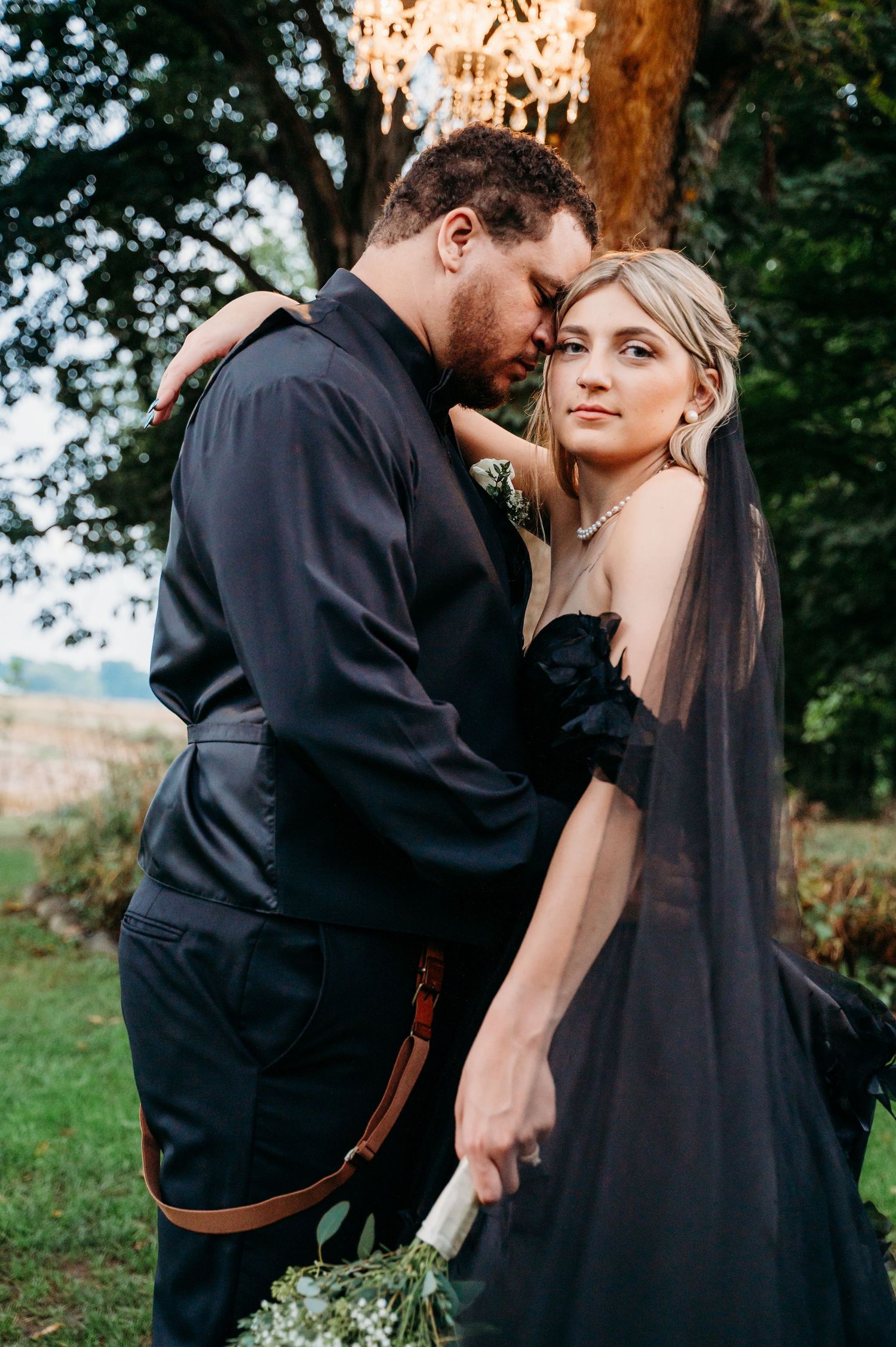 A romantic outdoor couple embrace while dressed in black, set against a natural green backdrop.