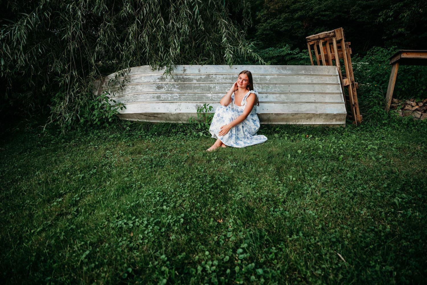 A figure in a light blue dress sits on rustic wooden steps surrounded by green grass and weeping willow branches.