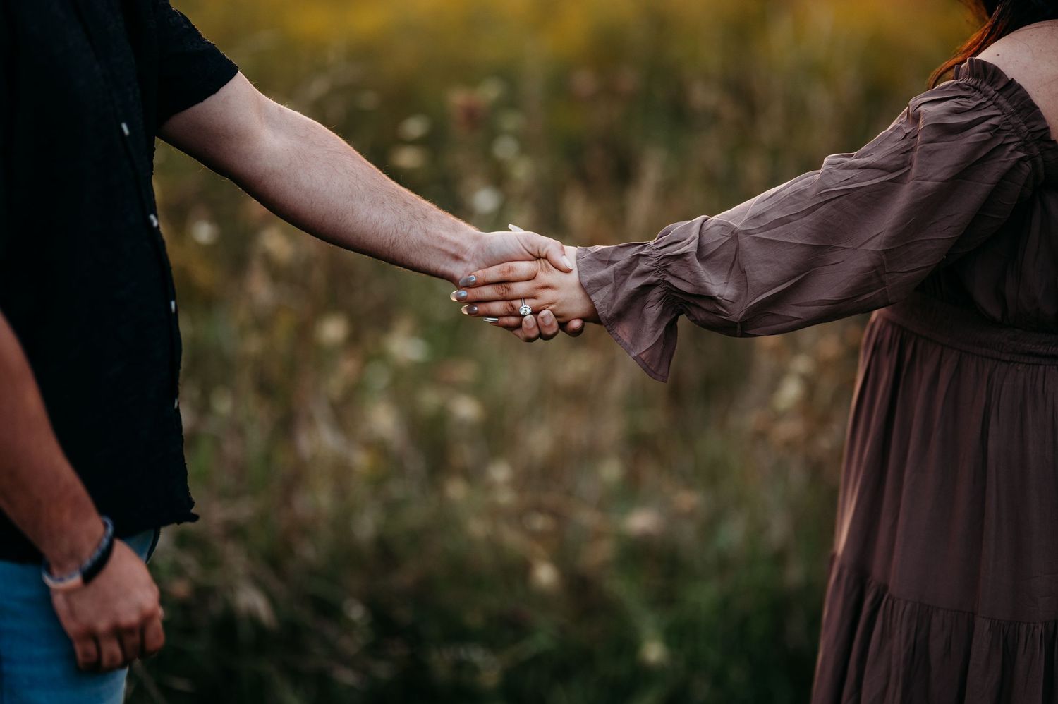 Two people hold hands in a romantic golden hour moment with brown and black clothing in a natural outdoor setting.
