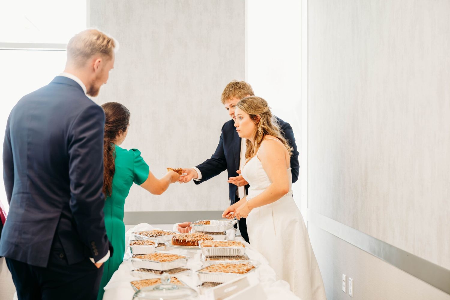 Bride and groom sharing cake at wedding dessert table with guests in elegant venue.
