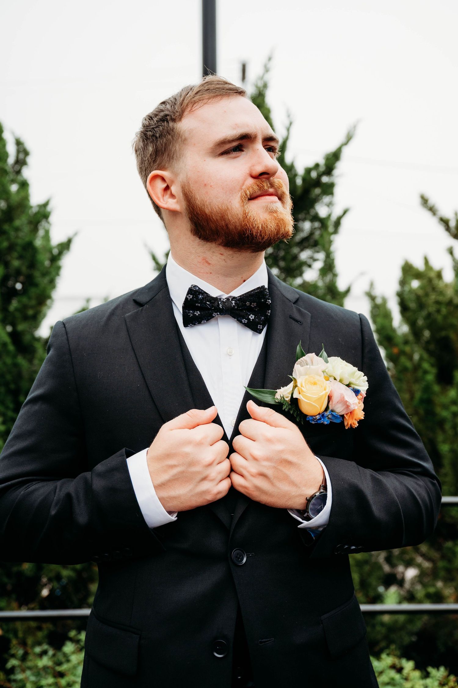 Handsome groom in black tuxedo with colorful boutonniere adjusting jacket outdoors near green hedges.