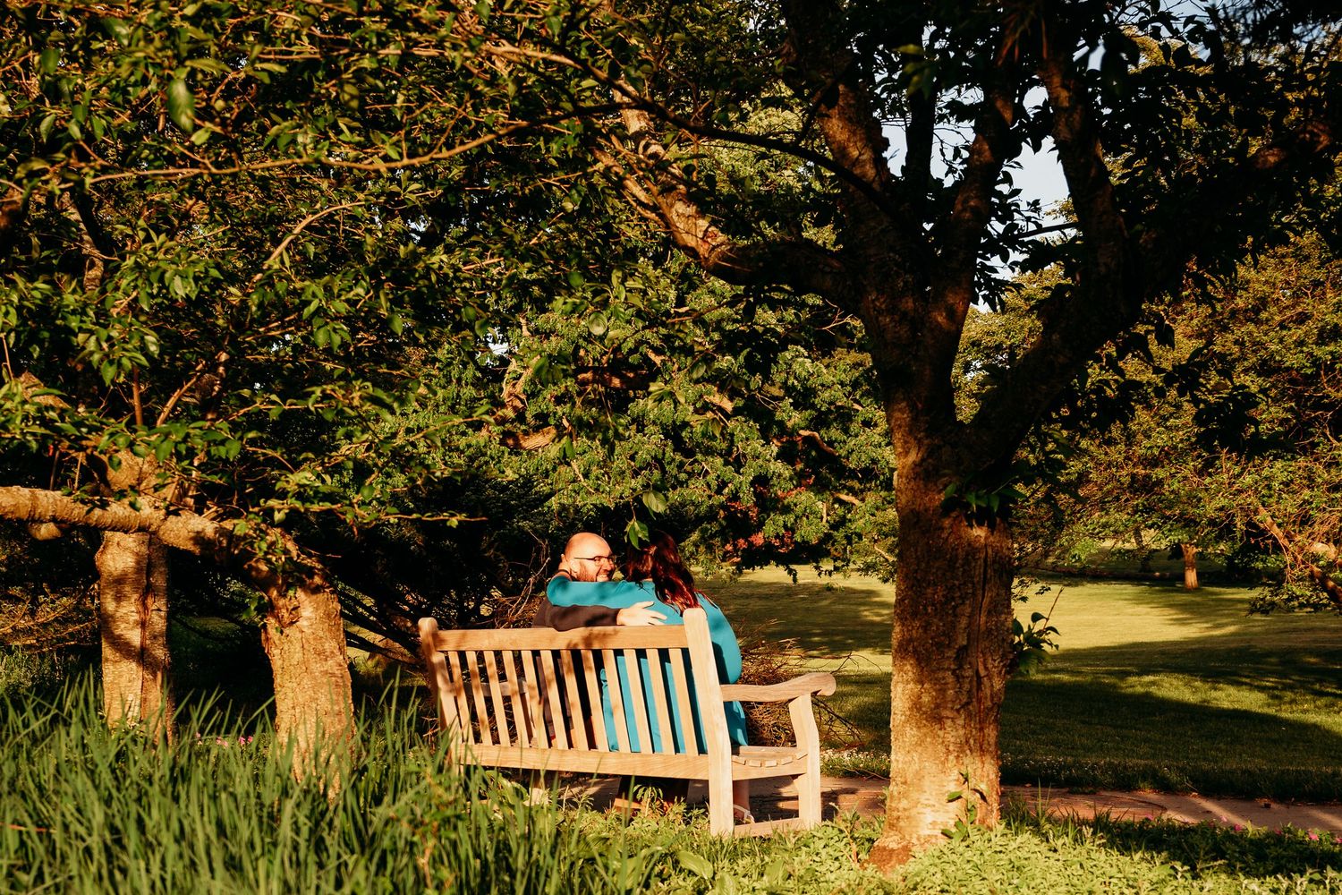 Person relaxing on a wooden bench under lush green trees in a sunlit park garden during golden hour.