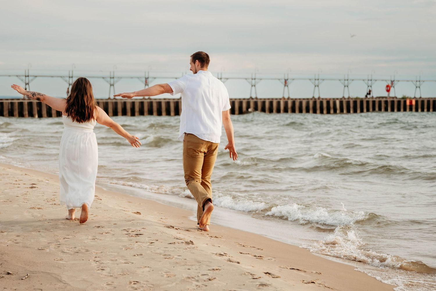 Couple walking hand in hand along a sandy beach near a pier, wearing white dress and khaki pants.